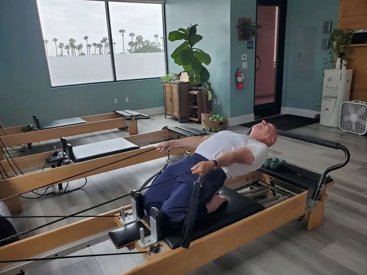 Person on a Pilates reformer in a studio, viewed from behind, with large windows and exercise equipment.