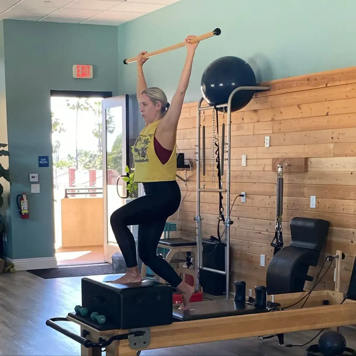 Person on a Pilates reformer in a studio, viewed from behind, with large windows and exercise equipment.
