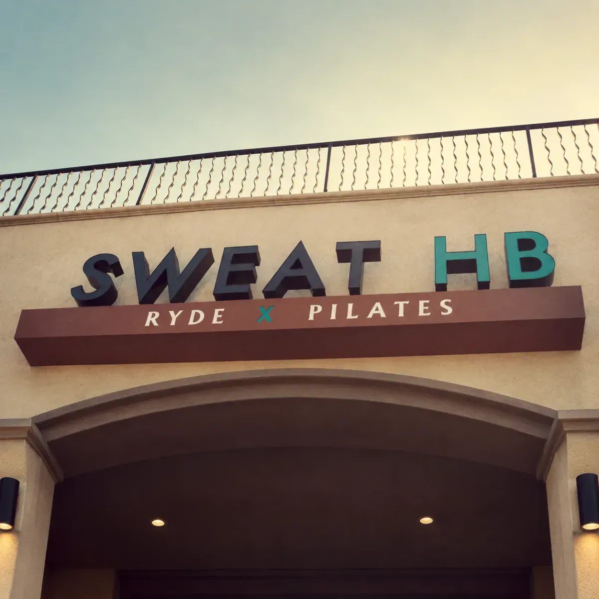 Storefront sign reading “SWEAT HB” with “RYDE x PILATES” below, on a tan building facade