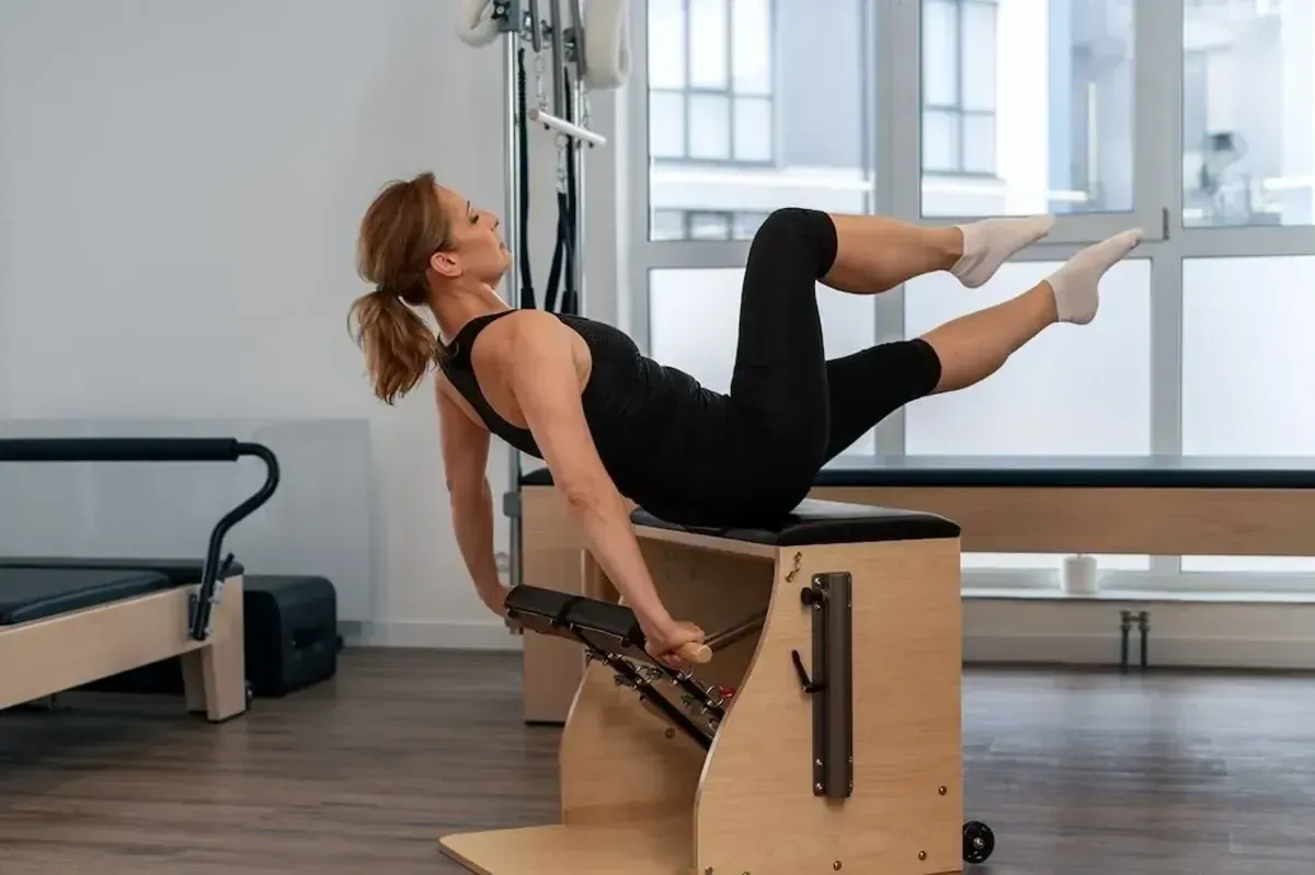 Person on a Pilates reformer in a studio, viewed from behind, with large windows and exercise equipment.