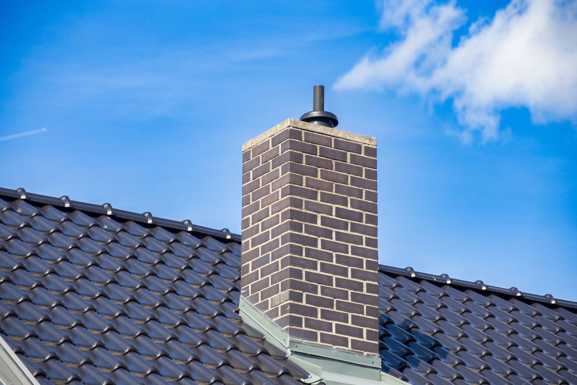 Brick chimney on a dark tiled roof against a blue sky with clouds