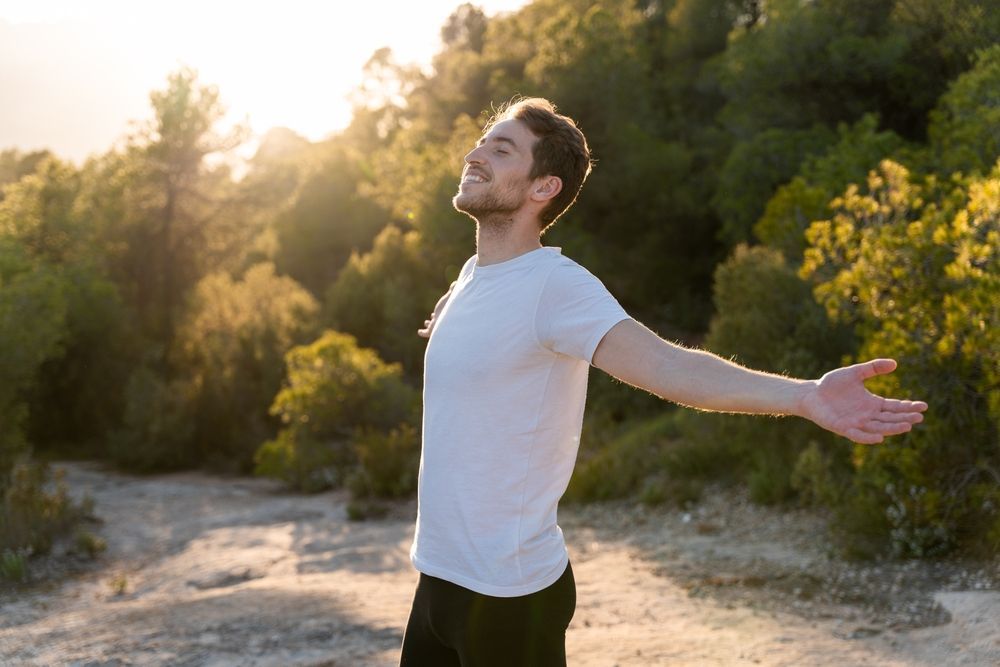 Person in a white shirt stands on a sunlit trail with arms outstretched, facing the trees.