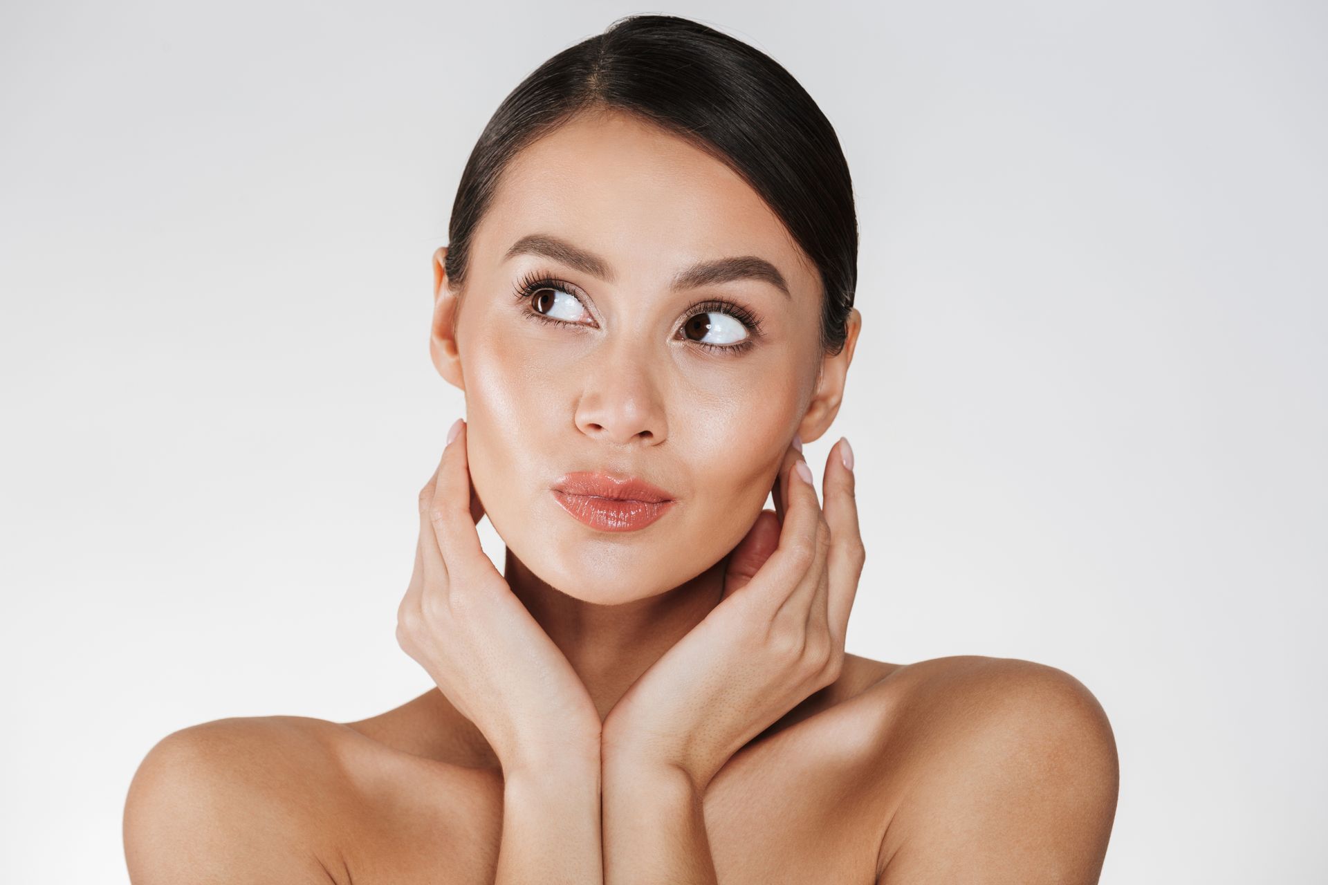 Woman with smooth skin, hands near face, looking upwards, light background.
