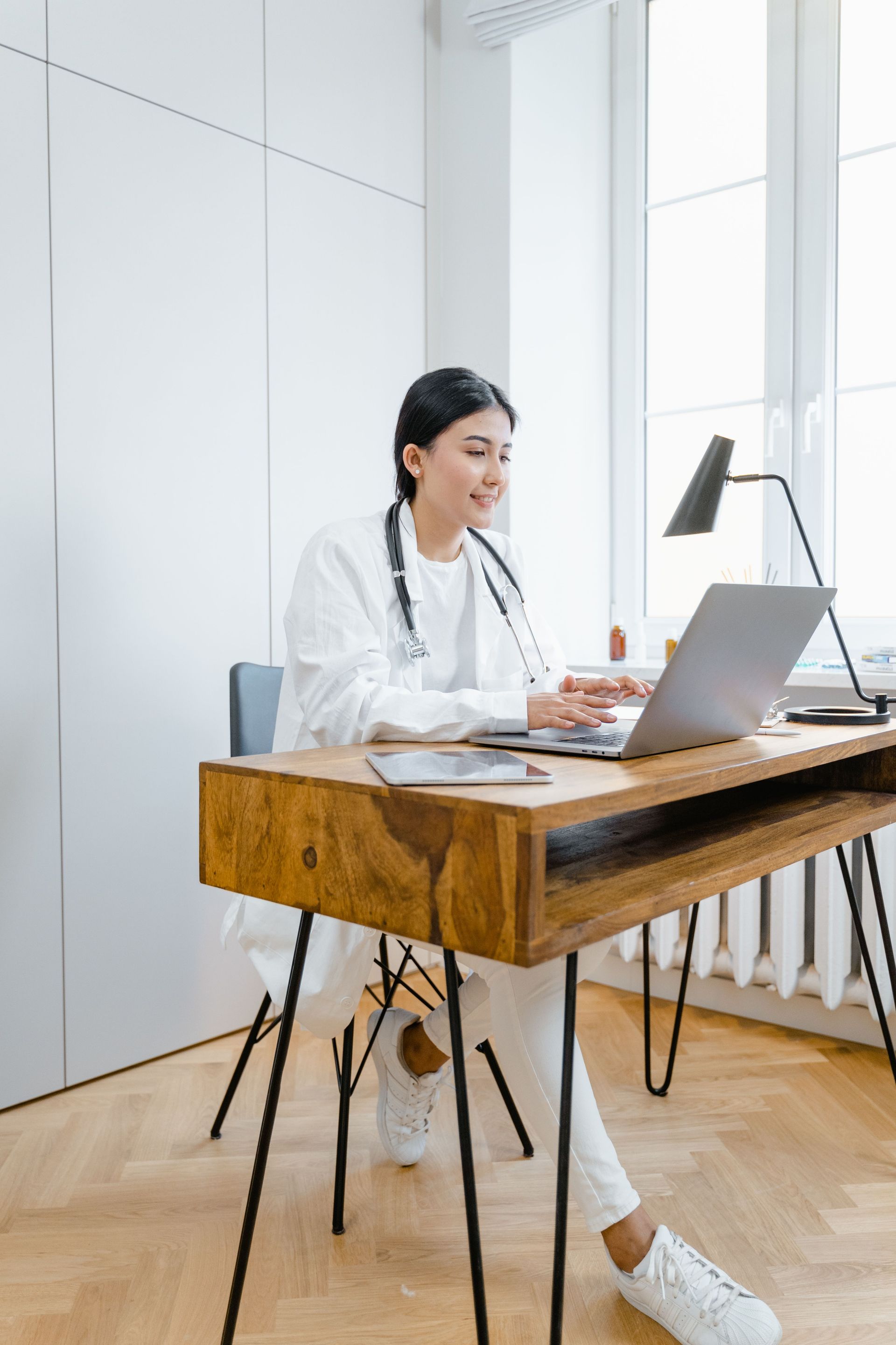 Woman working on a laptop at a wooden desk in a bright home office, with coffee and papers nearby