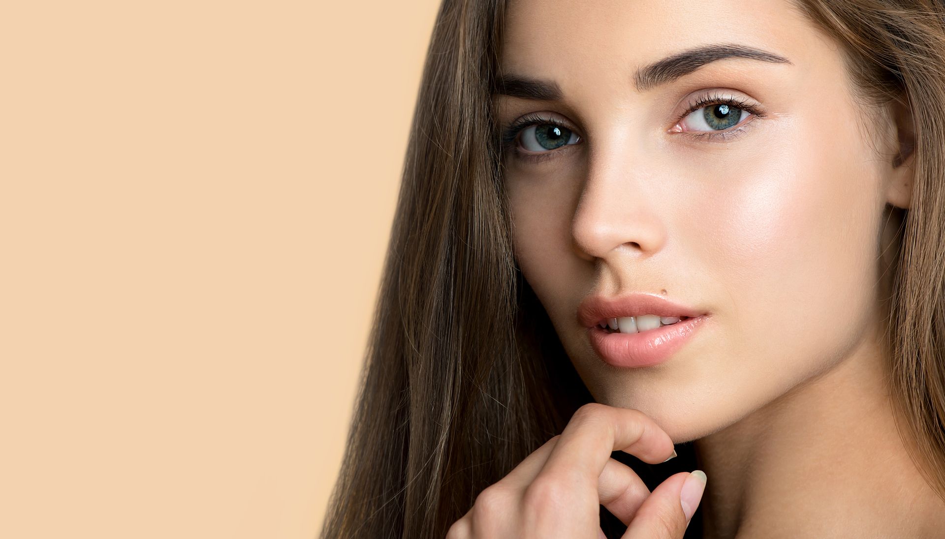 Woman with long brown hair, fair skin, and blue eyes, touching her chin, against a peach background.