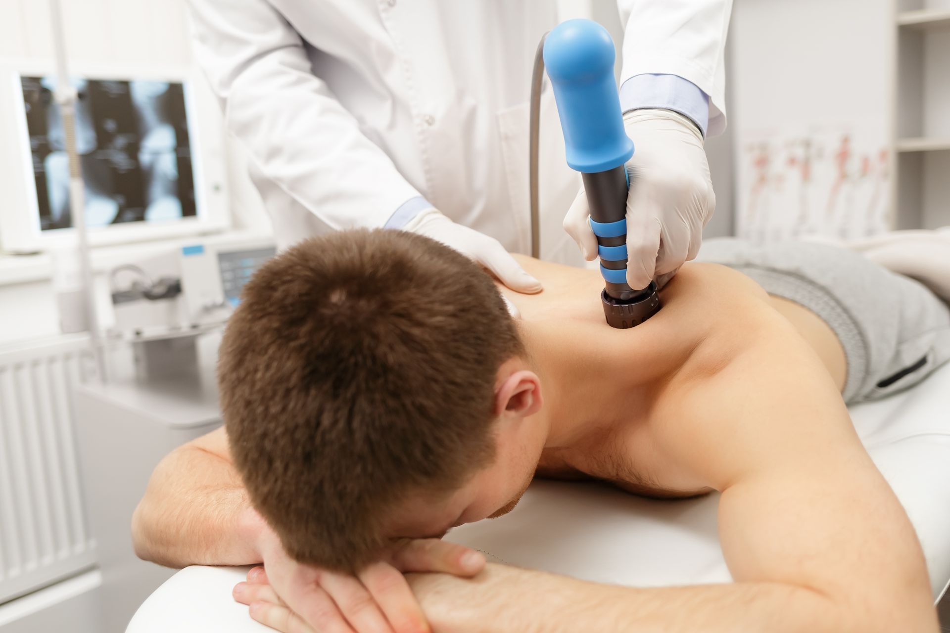 A doctor using a blue and black device on a patient's shoulder. The patient is lying face down. In a medical setting.