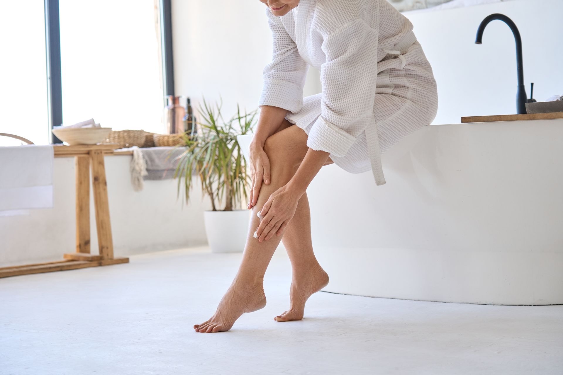 Woman in a white robe rubbing her lower leg in a bright bathroom or spa setting