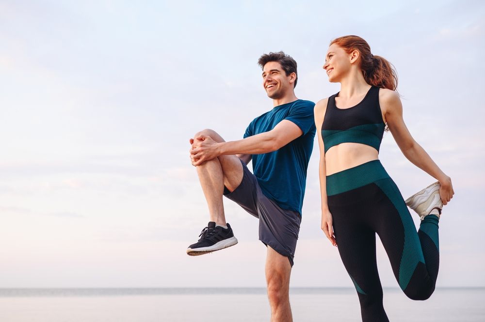 Two people in athletic wear stretching outdoors at sunrise near the beach