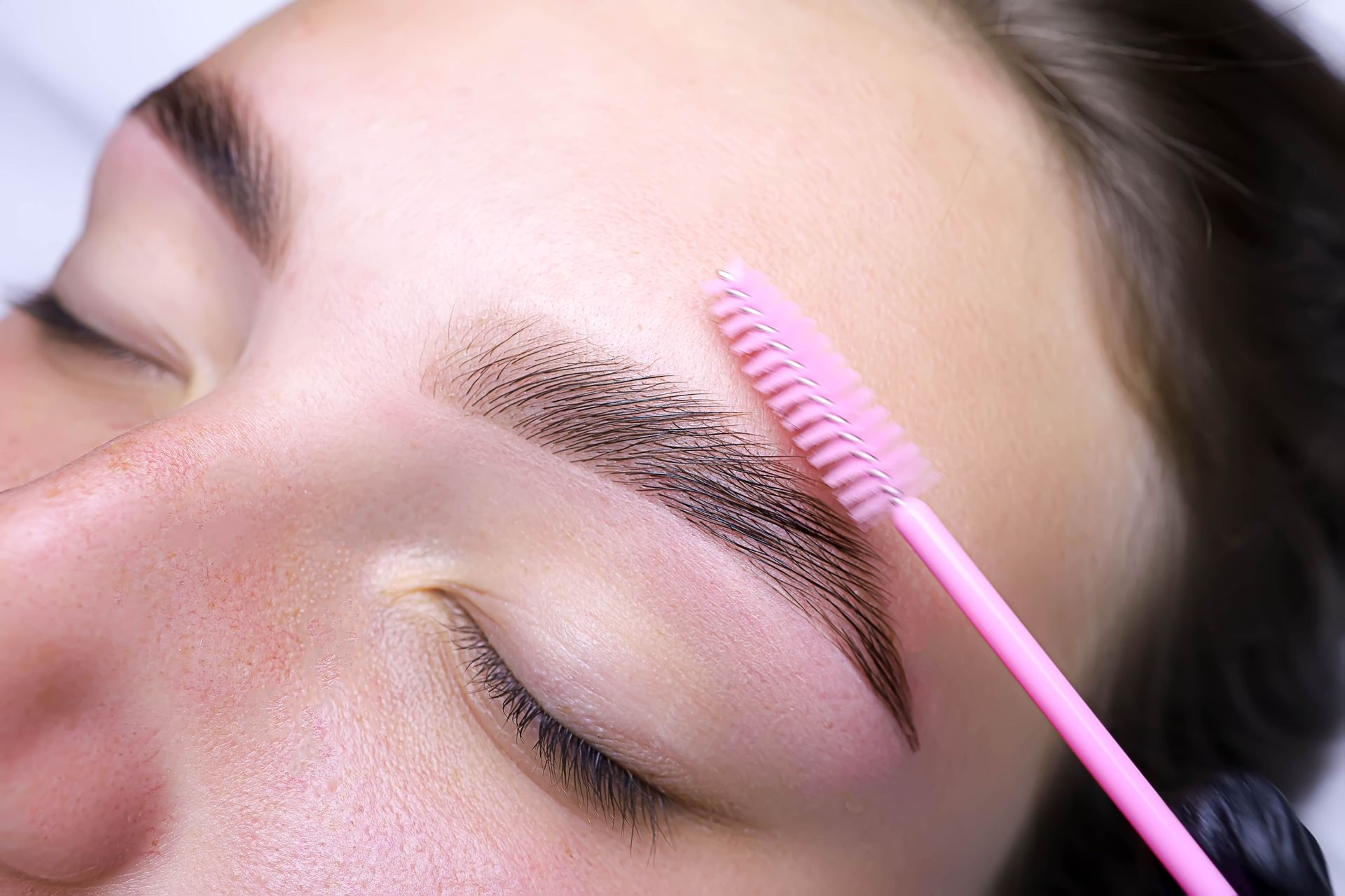 Close-up of a person with a groomed eyebrow being brushed upwards with a pink spoolie.