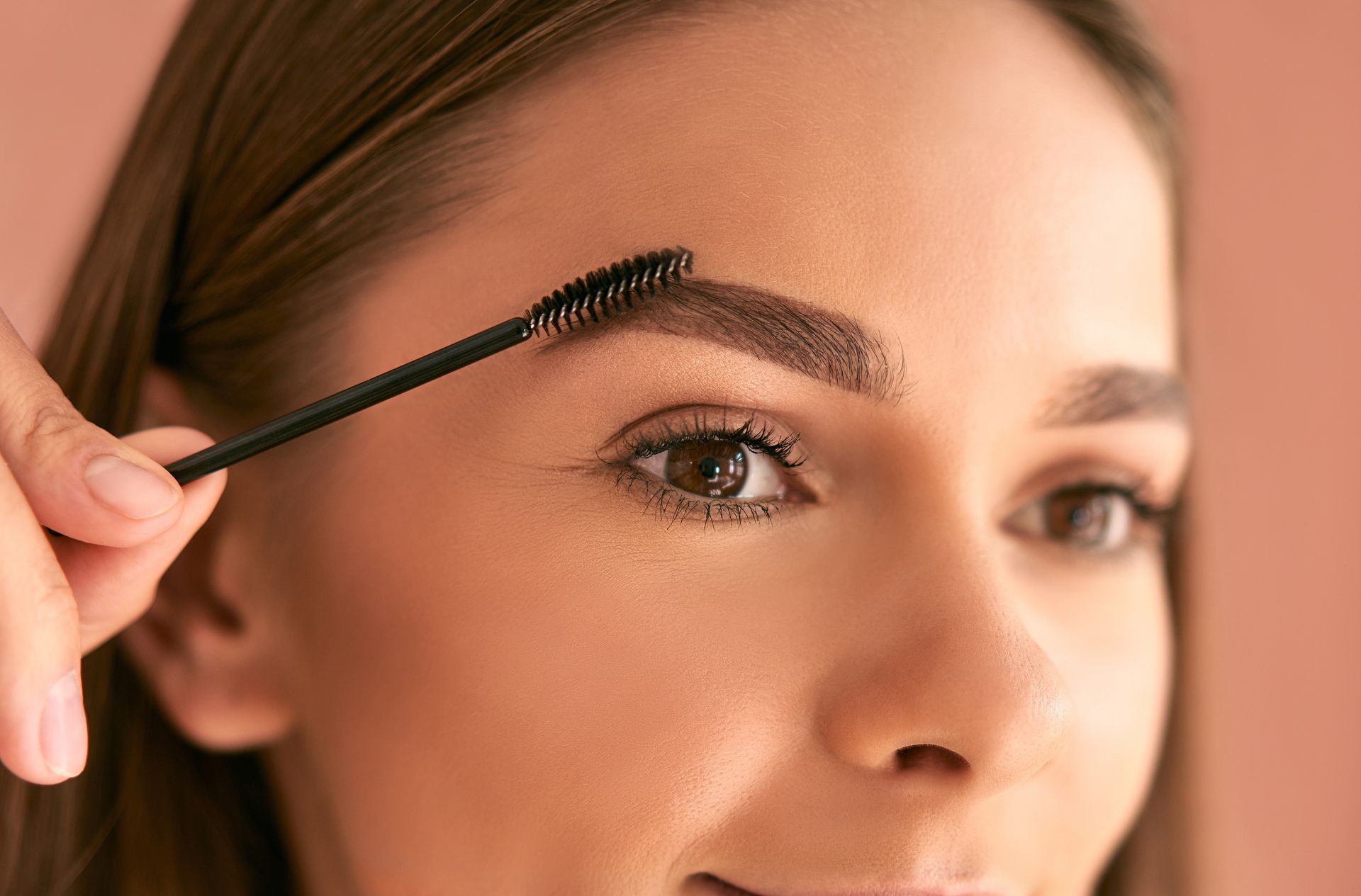 Woman brushing her eyebrows with a spoolie brush. She has brown eyebrows and eyes, and a peach background.