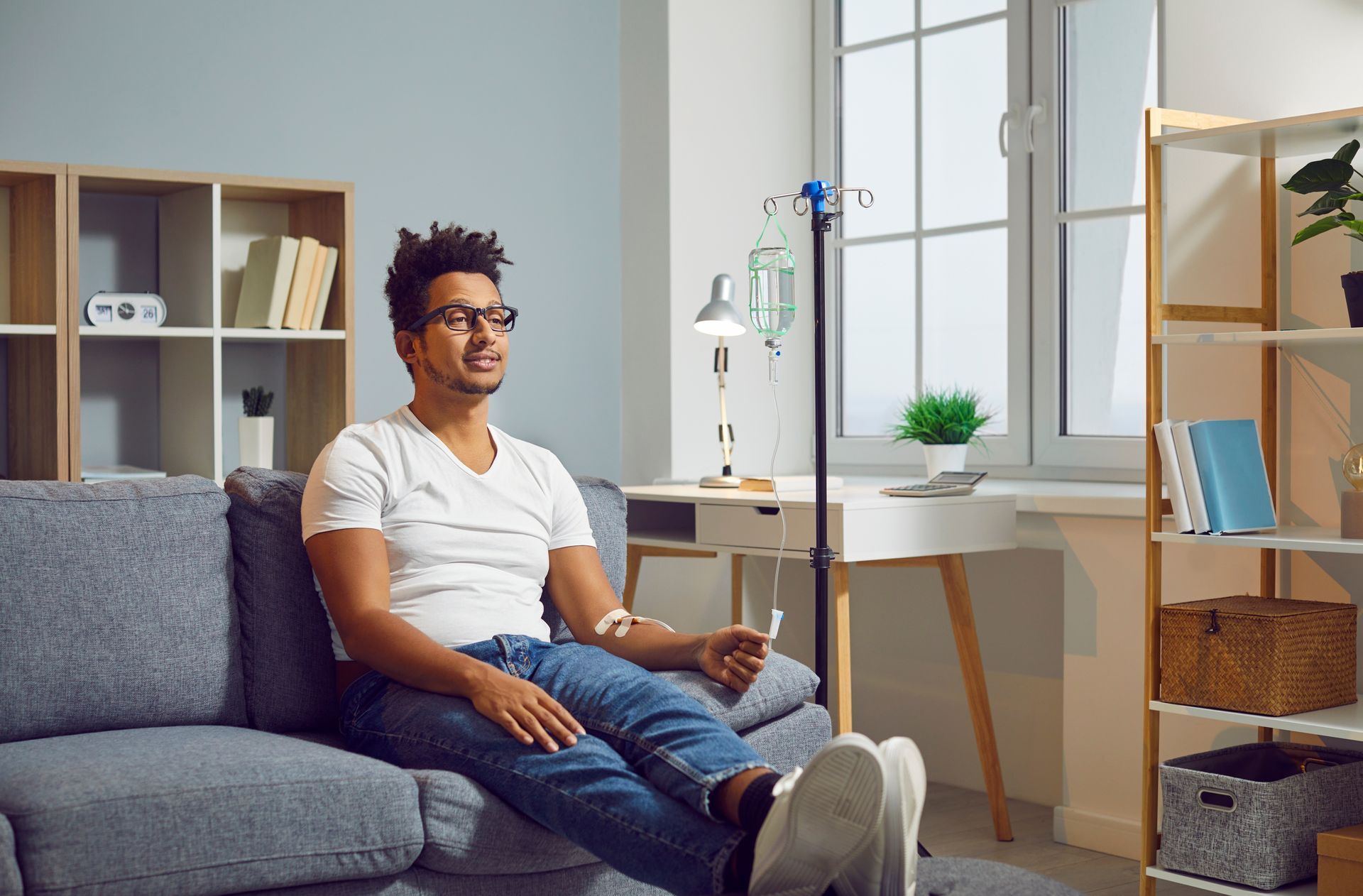 A Black man with glasses receives an IV infusion while relaxing on a sofa in a living room.