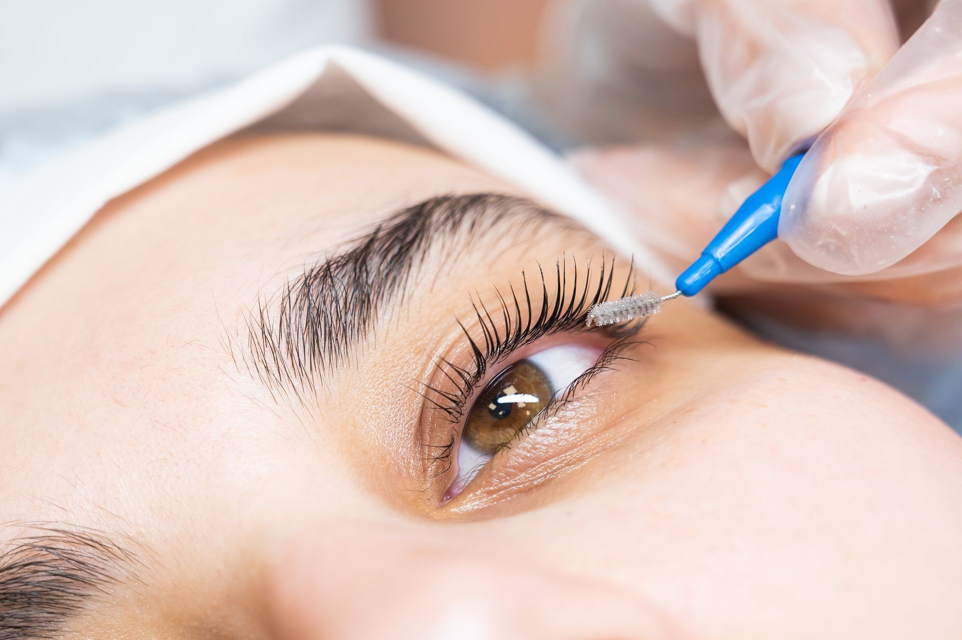 Close-up of a person receiving a lash lift. A gloved hand applies a product to curled eyelashes in a salon setting.