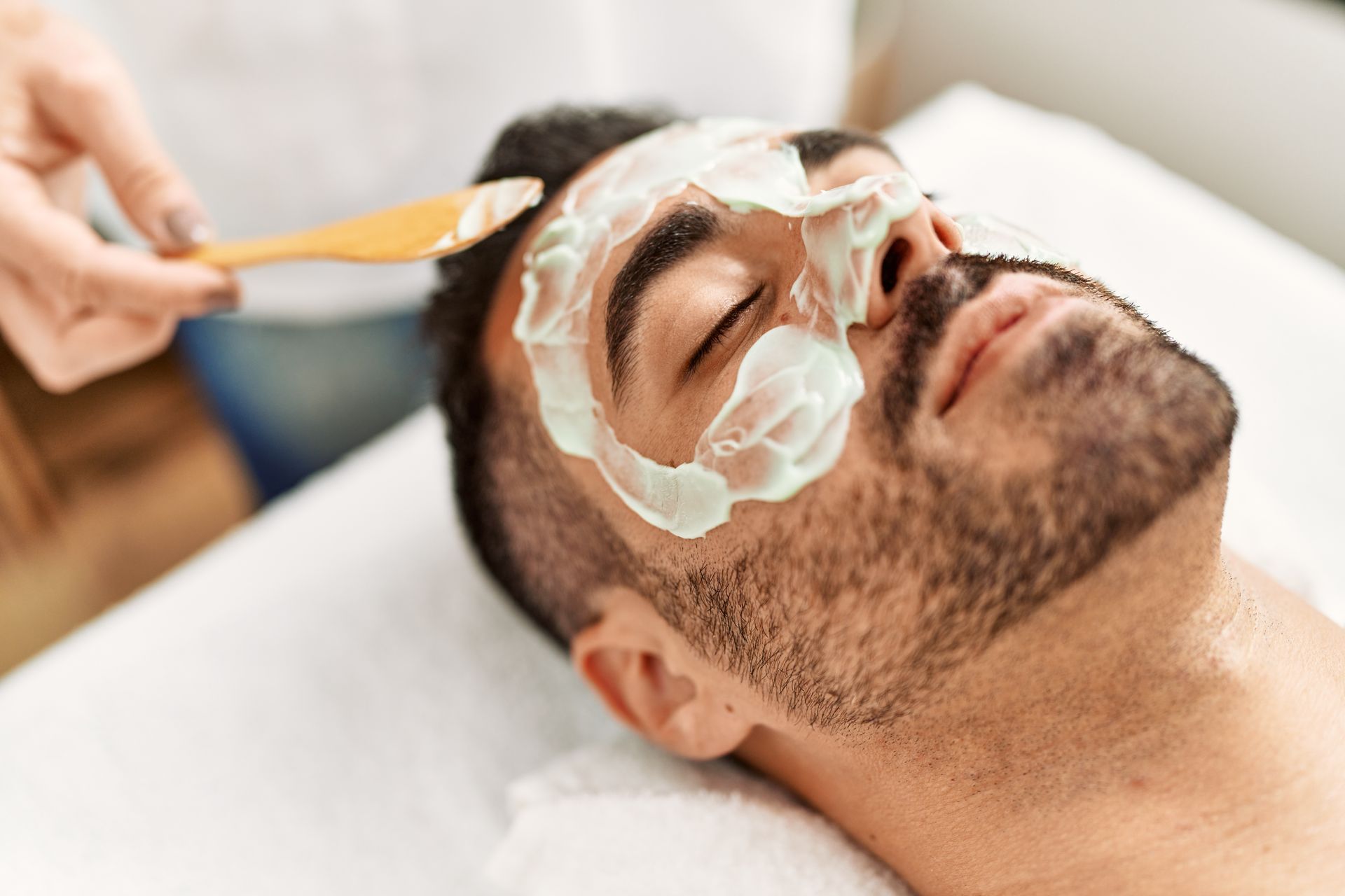 Man receiving facial treatment at a spa. A person applies a white mask to his face with a wooden spatula.