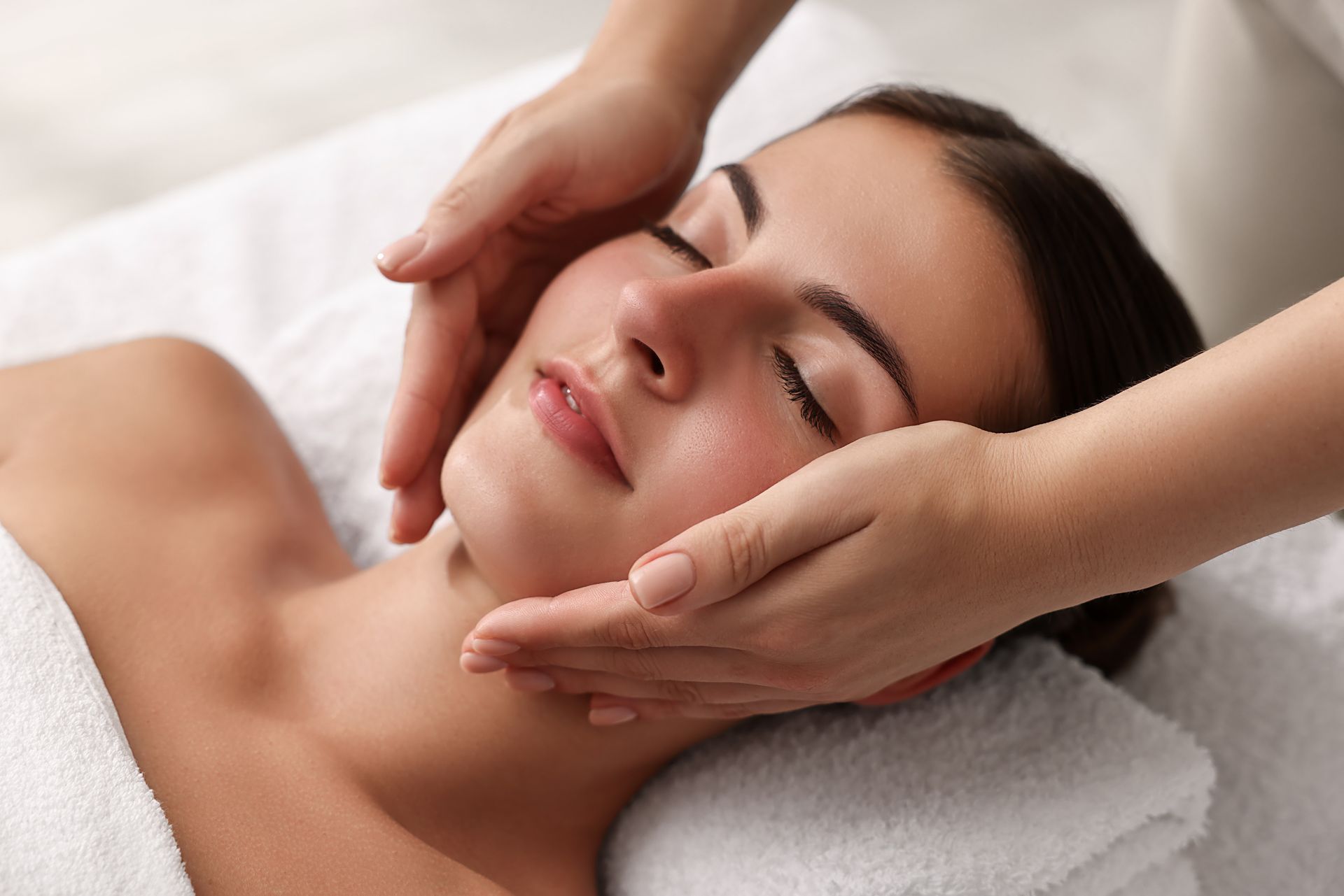 Woman receiving a facial massage. She is lying on a white towel with her eyes closed while two hands gently cradle her face.