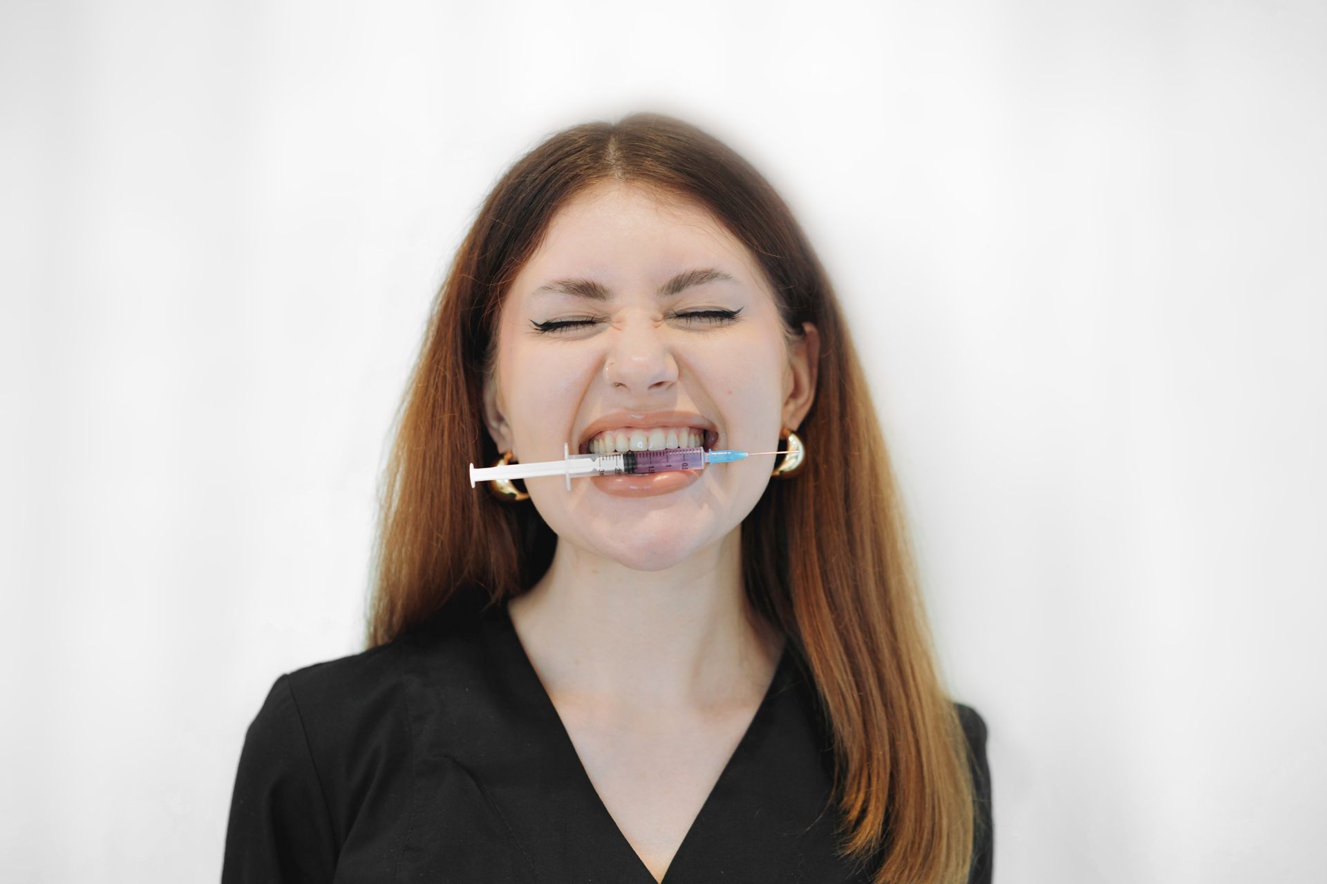 Woman with syringe in mouth, eyes closed, grimacing. Black top, gold earrings, white background.