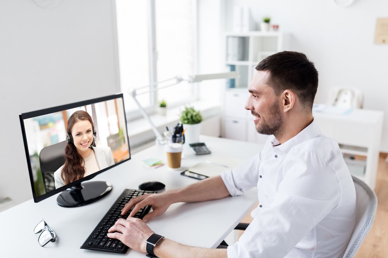 Man at a desk video chatting on a monitor, typing on a keyboard in a bright office