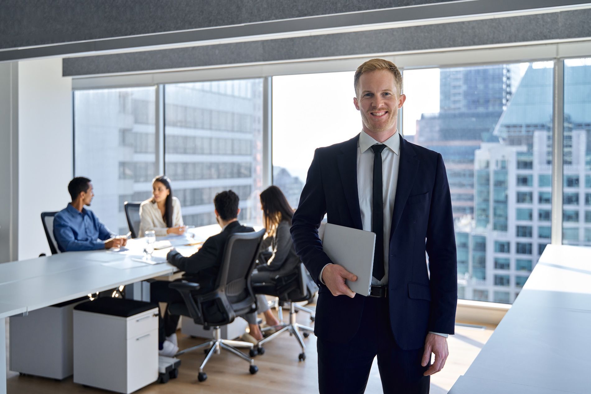 Business meeting in a modern office, with a man in a suit holding a laptop in the foreground and colleagues seated behind him.
