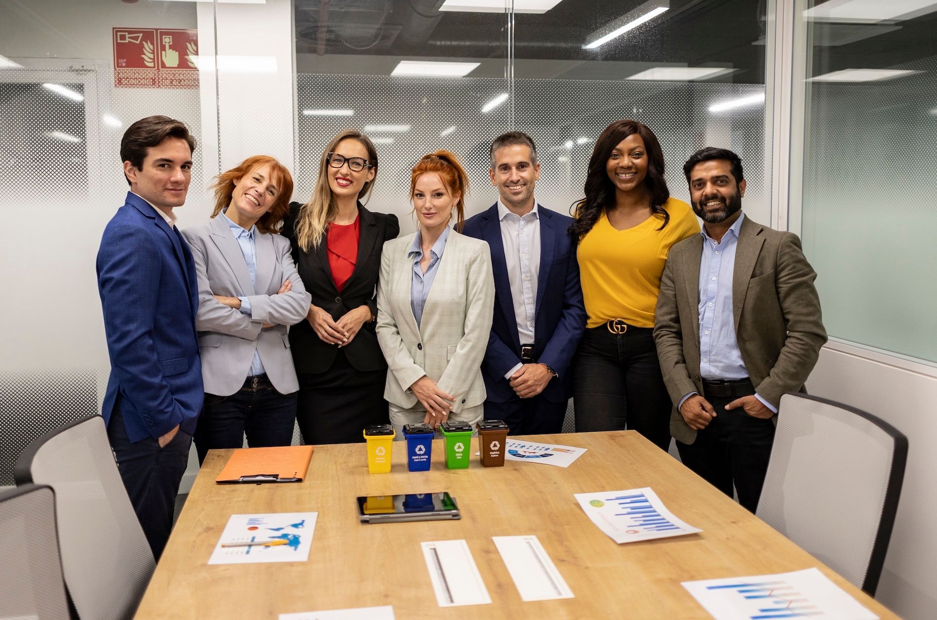 Six people standing around a conference table in a meeting room, smiling at the camera.