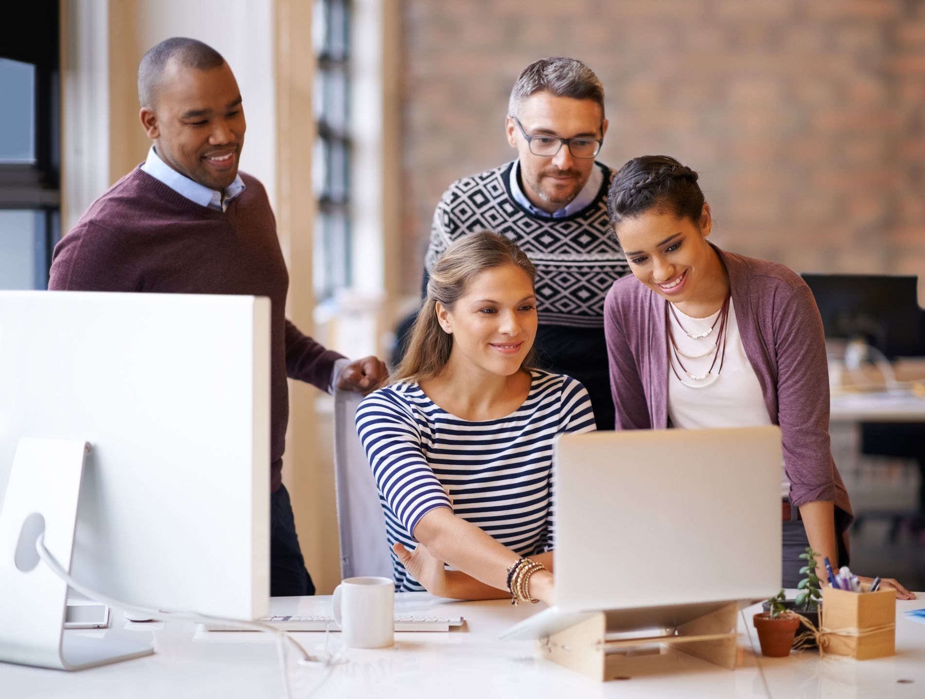 Four coworkers gather around a laptop in a bright office, smiling and collaborating on a project.