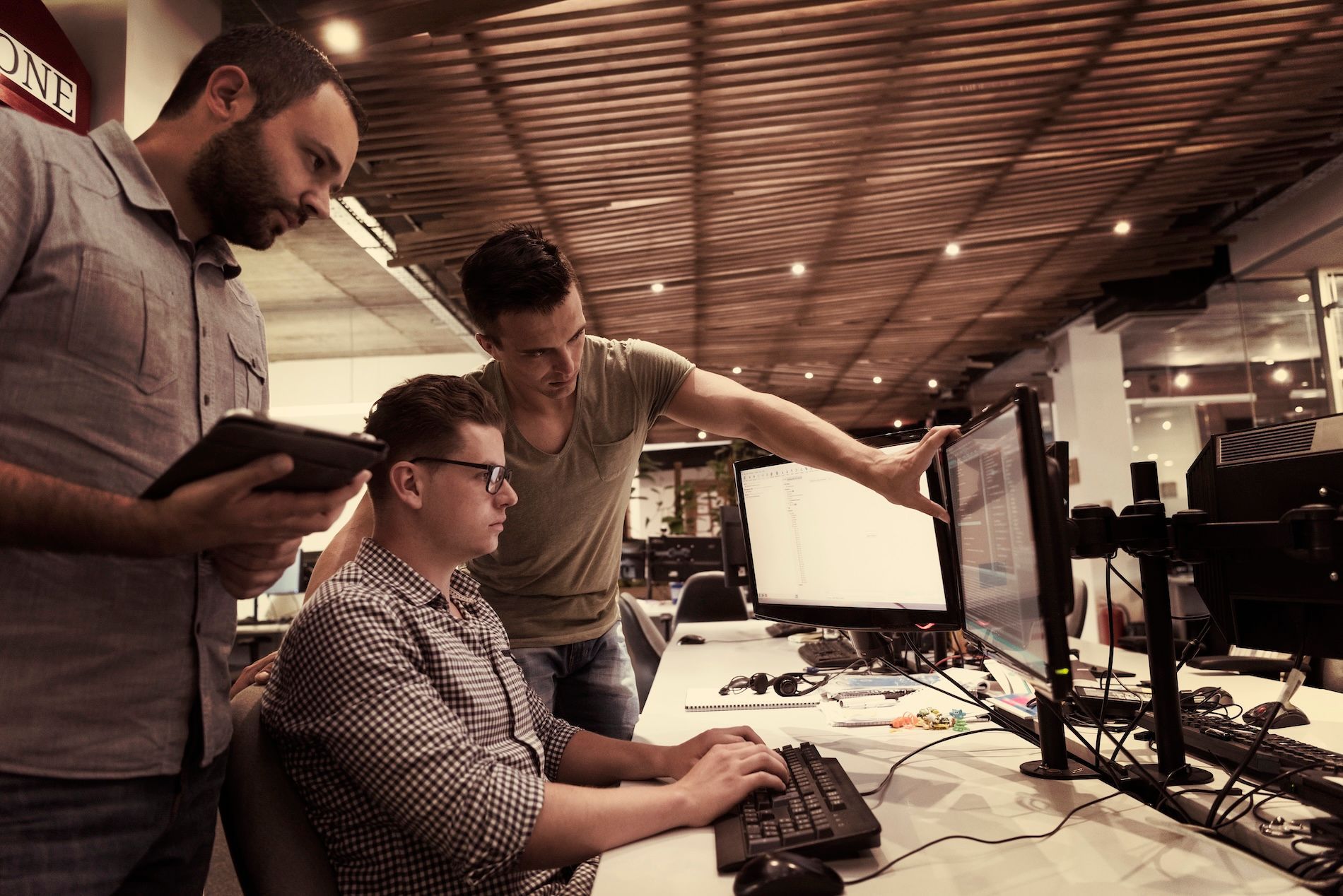 Three people collaborating at computer workstations in an office, one seated and two standing over monitors.