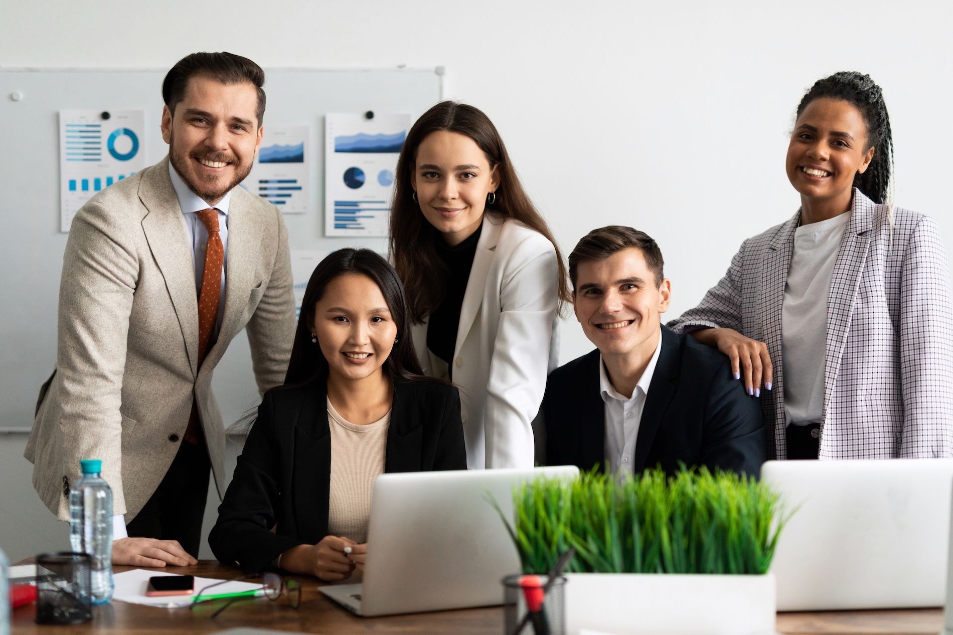 Five coworkers pose in a bright office, smiling around laptops and a desk with a small plant.