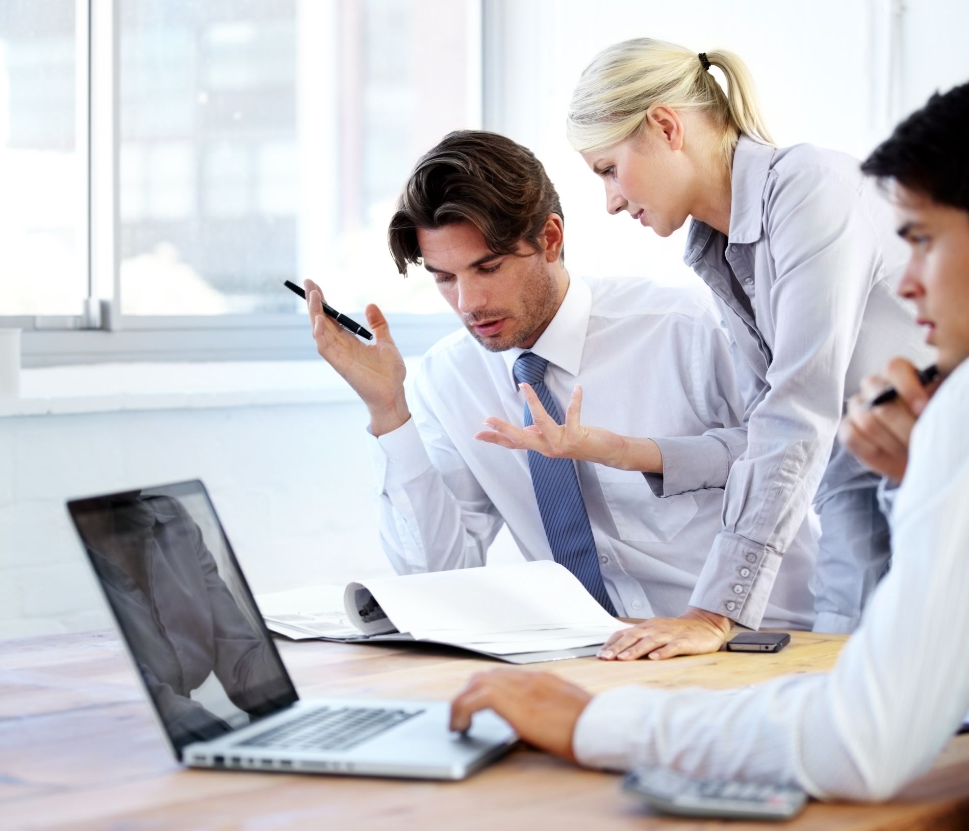 Office meeting with three coworkers gathered around a laptop and papers, discussing charts in a bright conference room