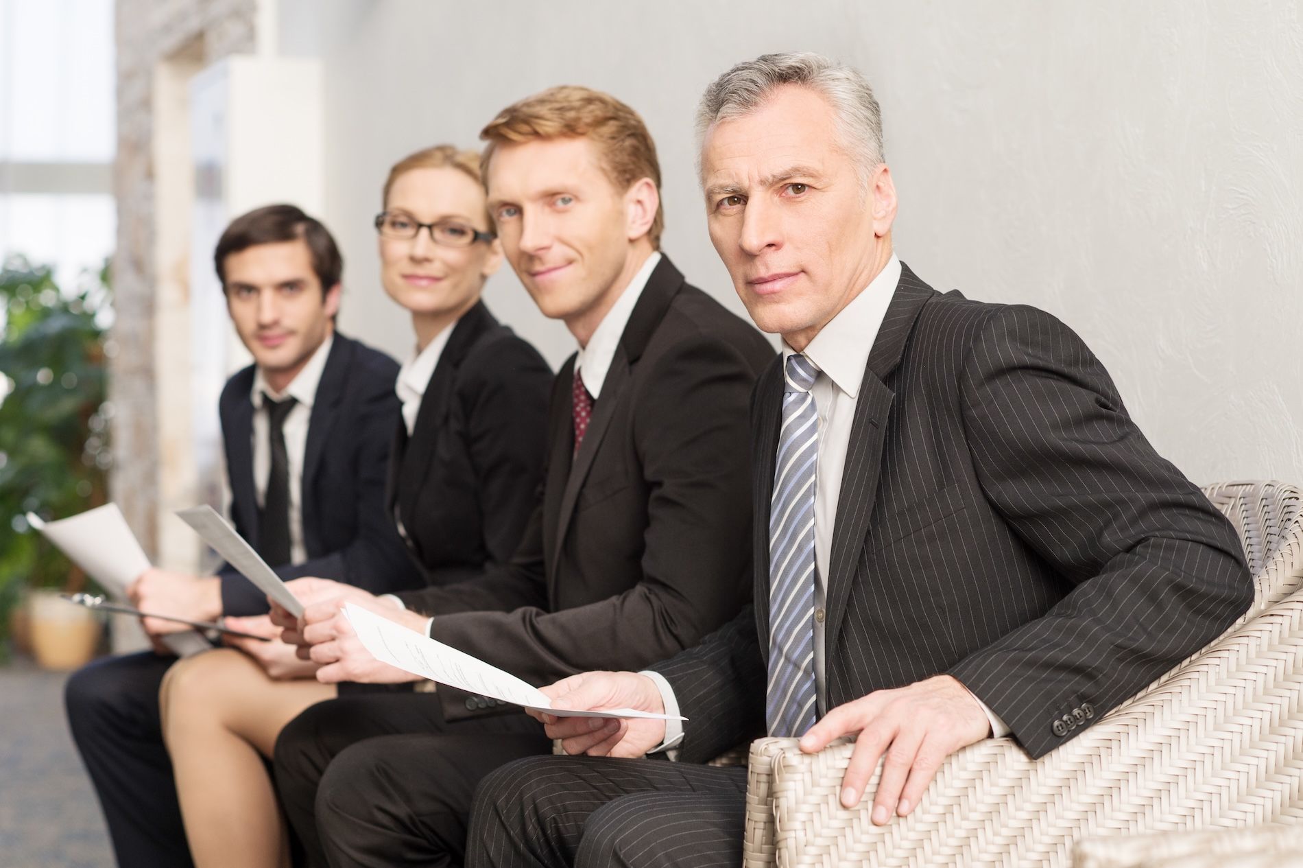 Four professionals in suits sitting on a sofa, holding papers in a bright office lobby.