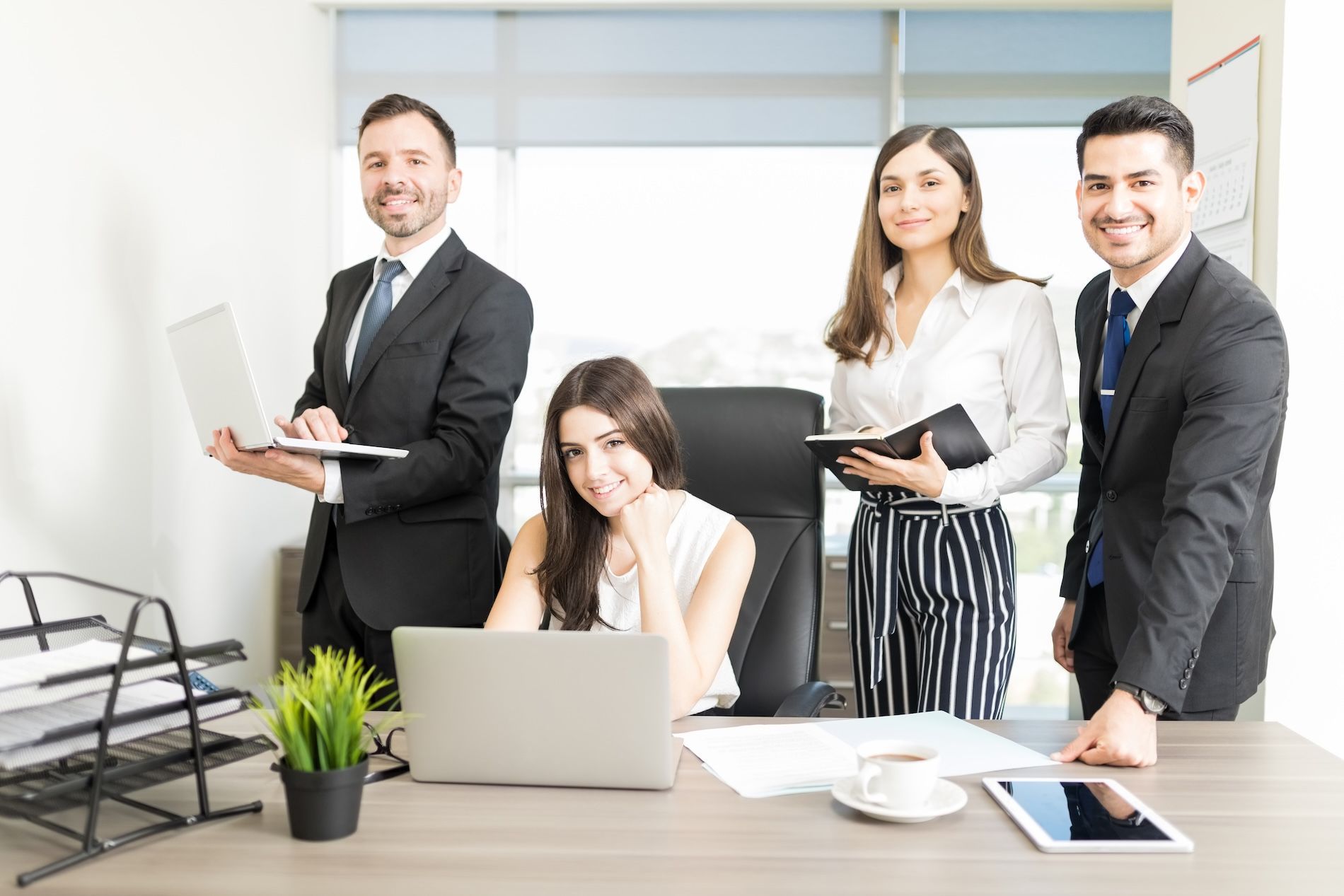 Four business professionals in an office, standing and sitting around a desk with laptops and documents.
