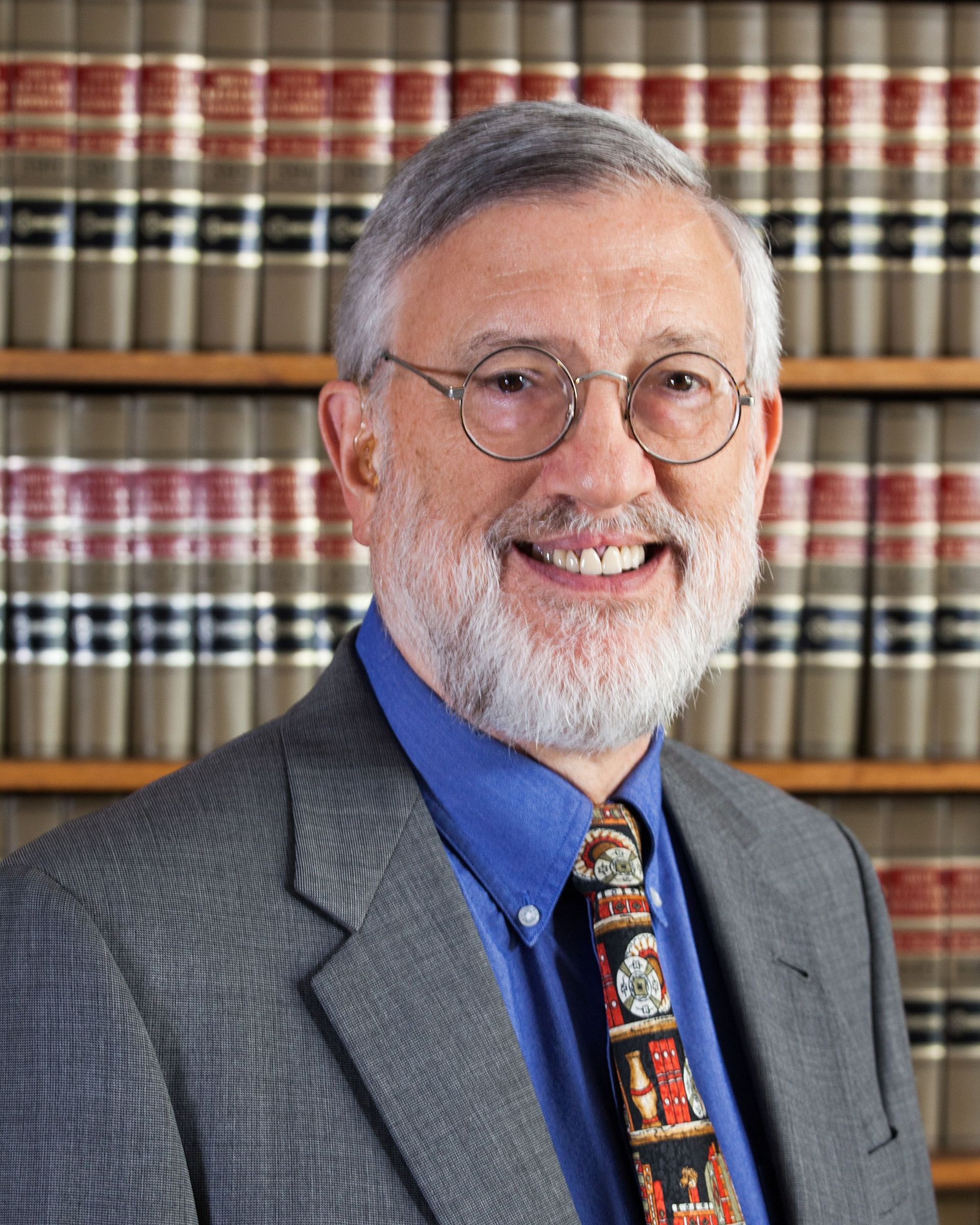 Smiling older man with glasses, grey hair and beard, wearing a blue shirt and patterned tie, in front of bookshelves.