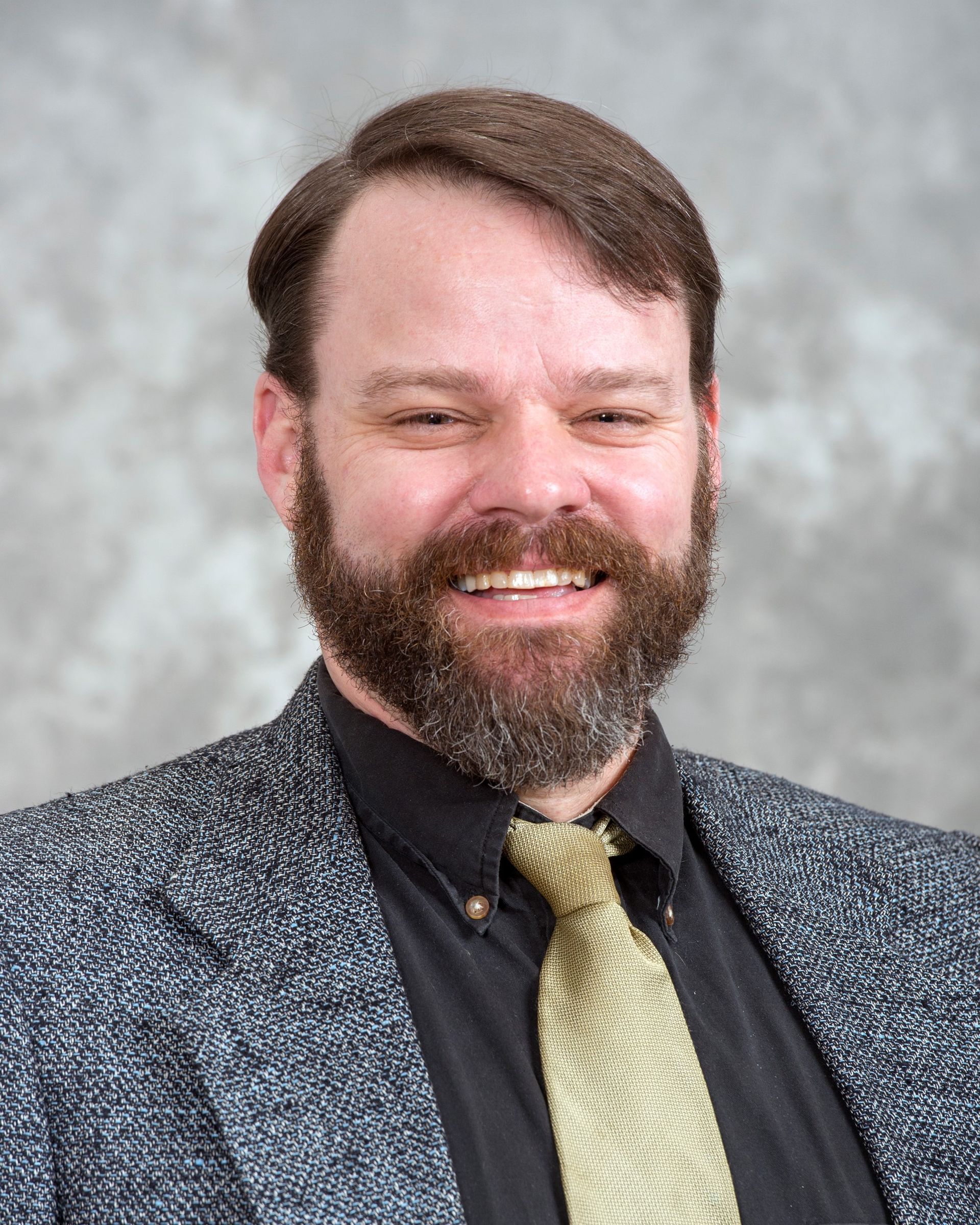 Man with a full beard, smiling, wearing a blazer and gold tie.