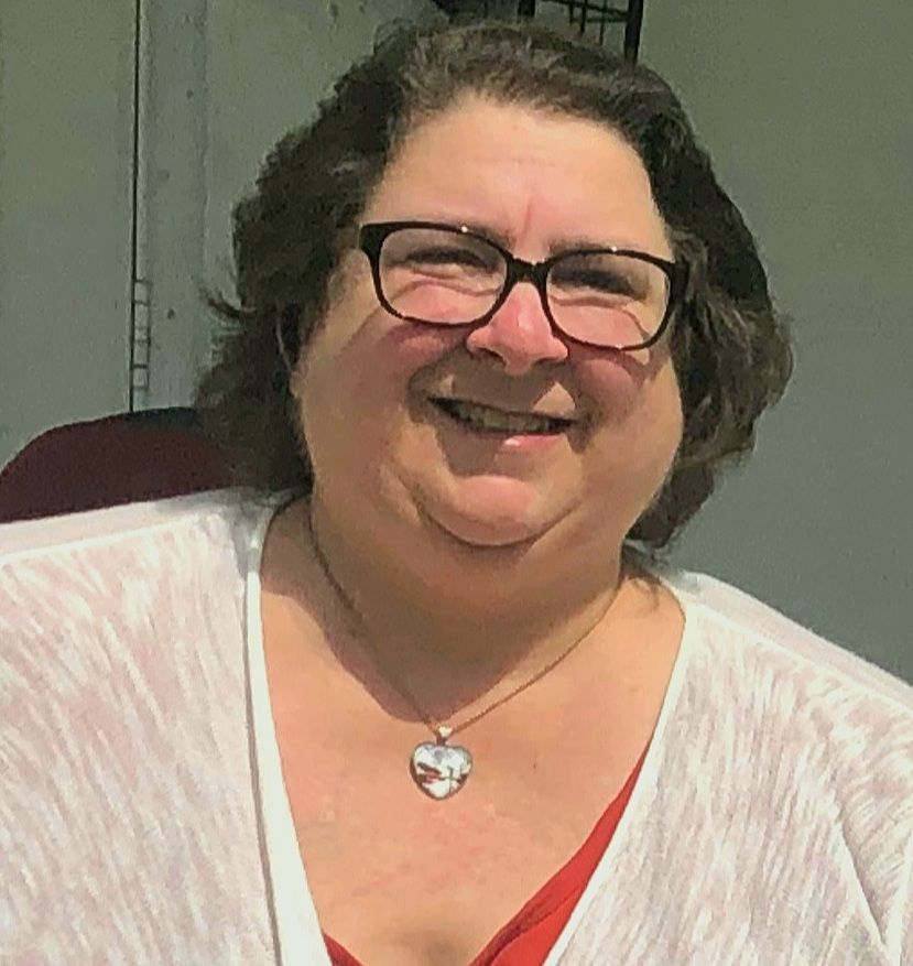 Woman wearing glasses and a heart pendant smiles, outdoors, dressed in light and dark tops.