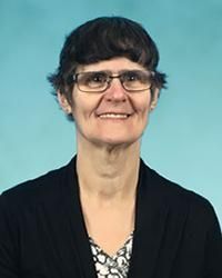 Woman with short dark hair and glasses, smiling in black jacket against blue background.