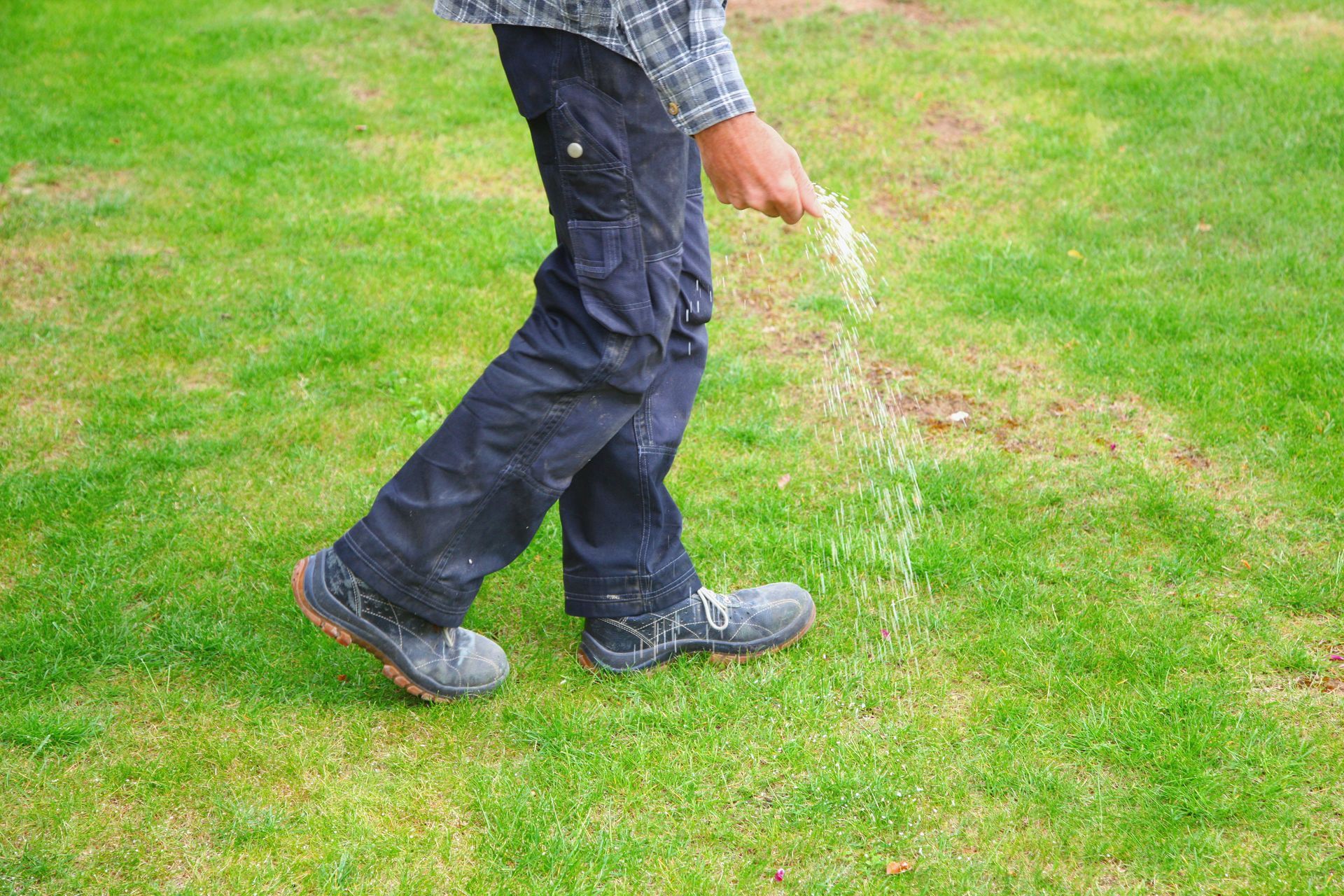 Person walking on grass, wearing dark pants and boots, holding a green watering can