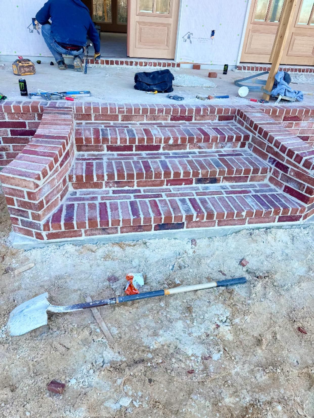 A worker kneels on a porch behind newly laid brick steps in front of a house, with a shovel resting on the sandy ground.