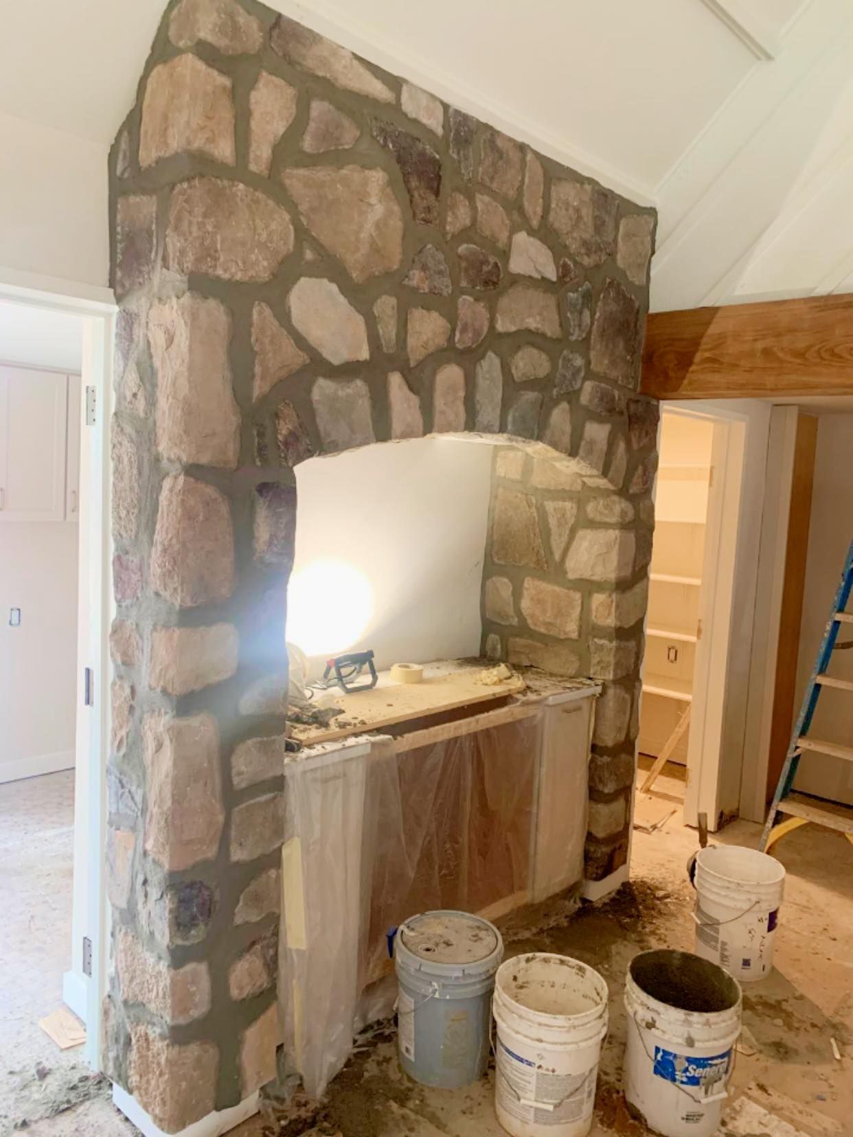 A stone-covered archway under construction in a room, with plastic sheeting covering cabinets and buckets on the floor.