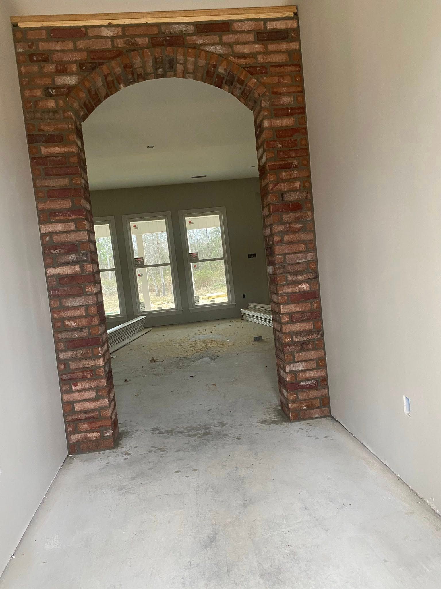 A rustic brick archway frame in an unfinished room, showing windows and a debris-covered floor beyond.