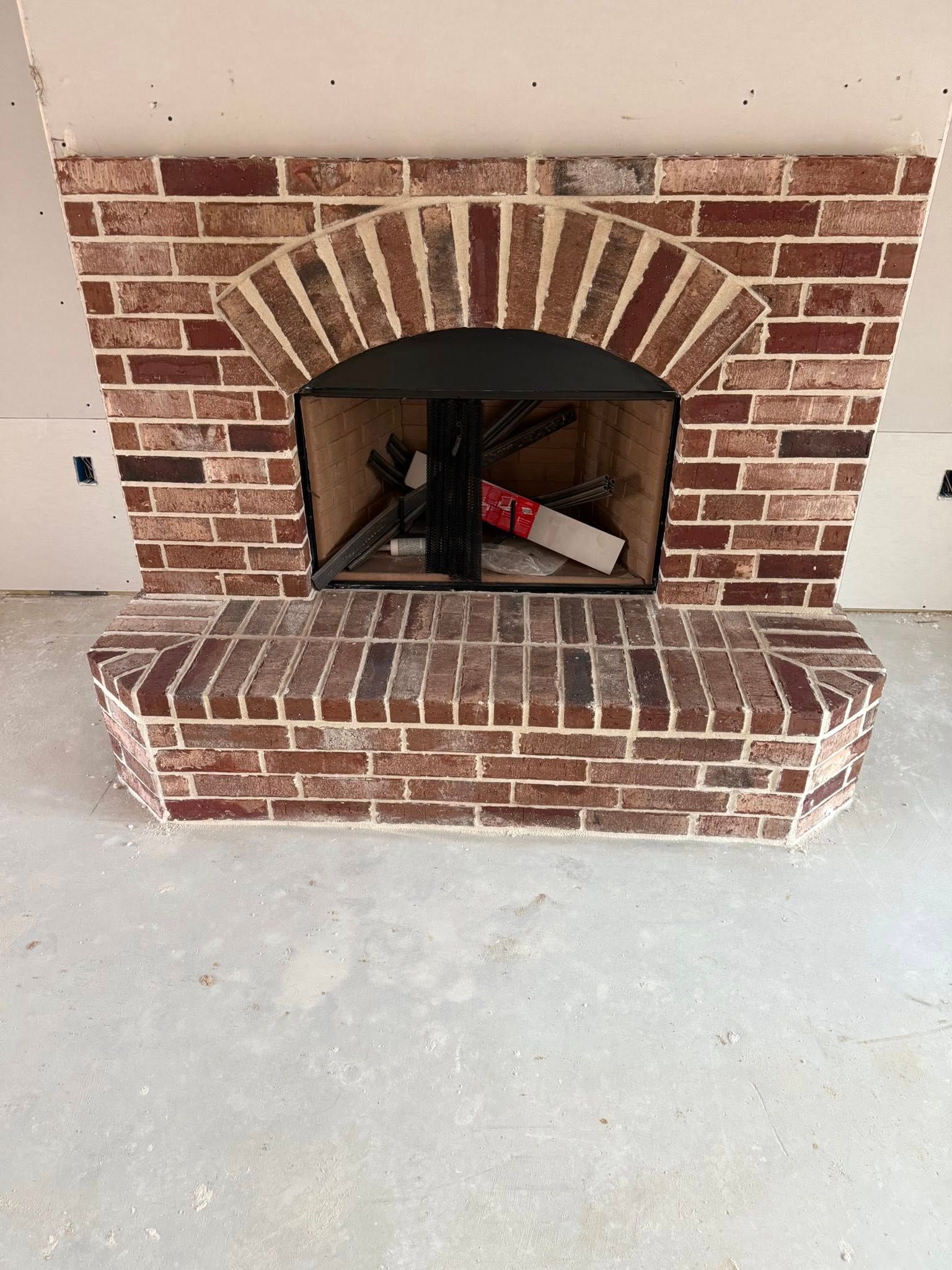 A brick fireplace with an arched opening and a raised brick hearth, set against unpainted drywall in a room under repair.