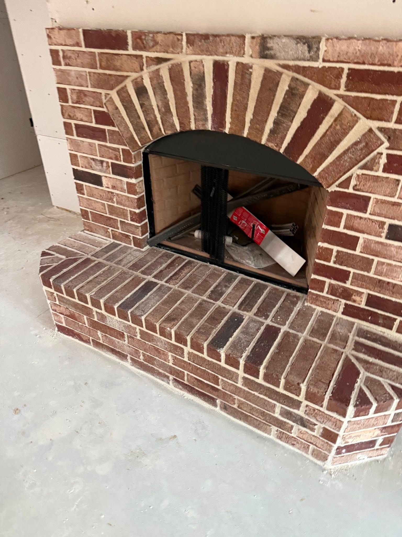 A red brick fireplace with an arched opening and a raised brick hearth in an unfinished room.