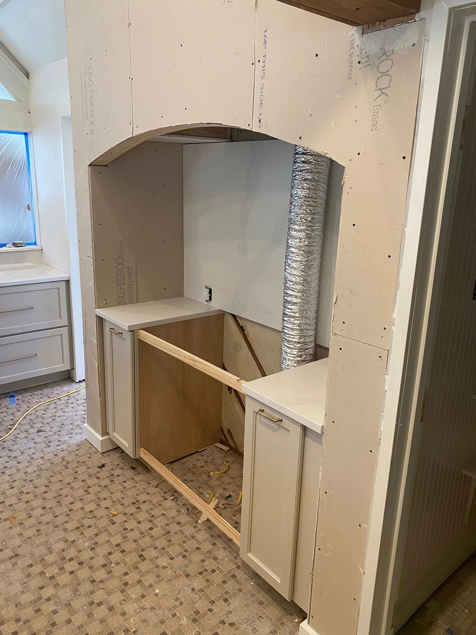 A kitchen renovation featuring a custom arched range hood enclosure above unfinished cabinetry and a metallic vent pipe.