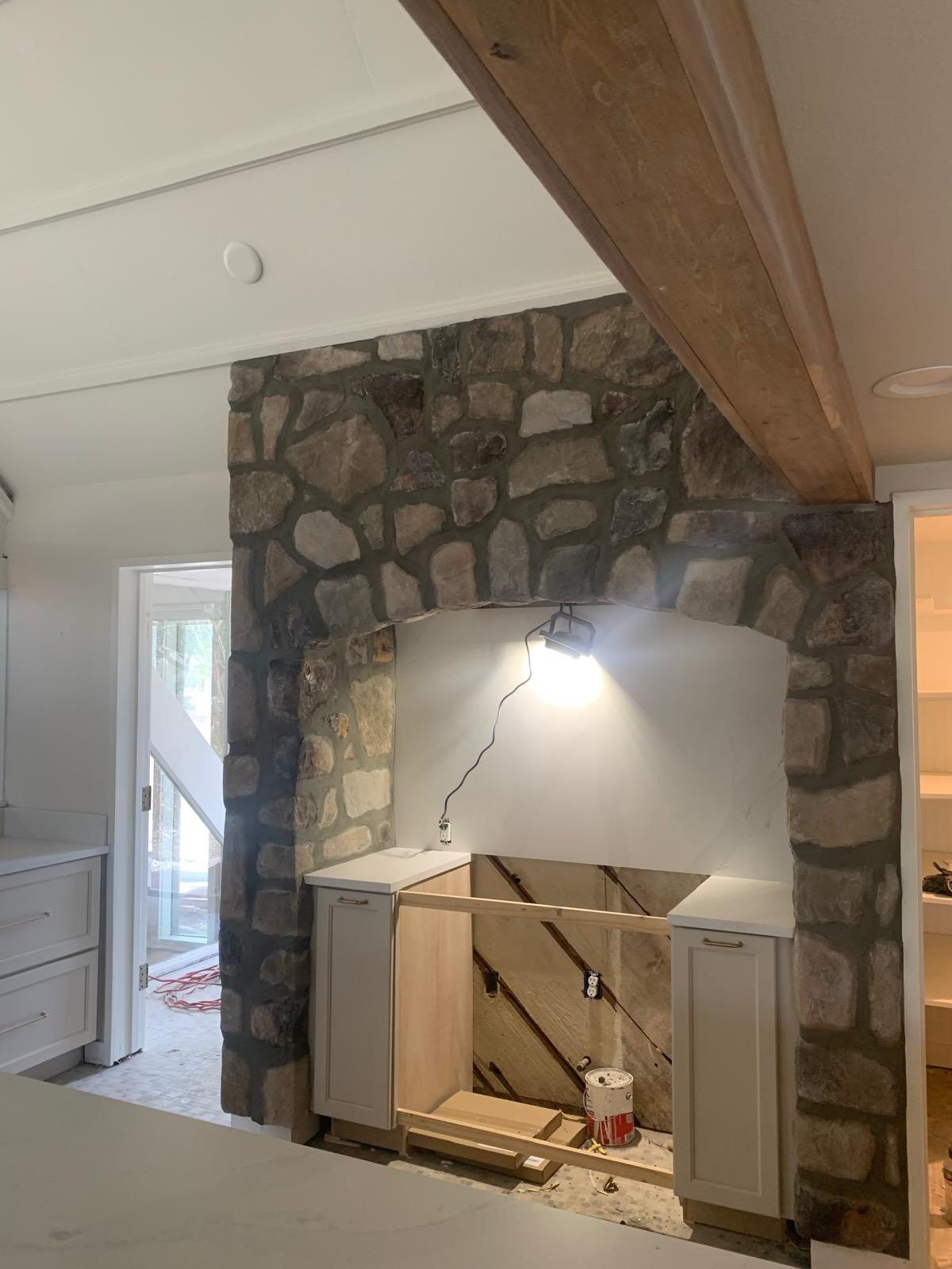 A stone archway surrounds an unfinished kitchen stove alcove with white cabinets and a bare wall in a bright room.