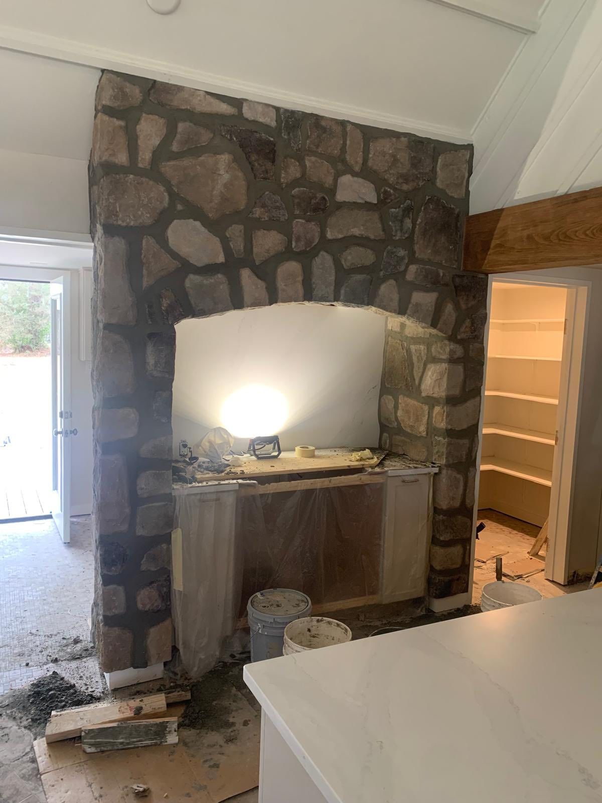 A stone-clad arched fireplace structure under construction in a room, with building materials and buckets on the floor.