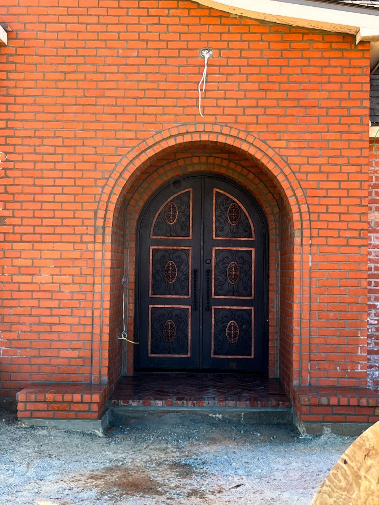 A red brick arched entryway features ornate, dark double doors with copper-colored trim and hardware.
