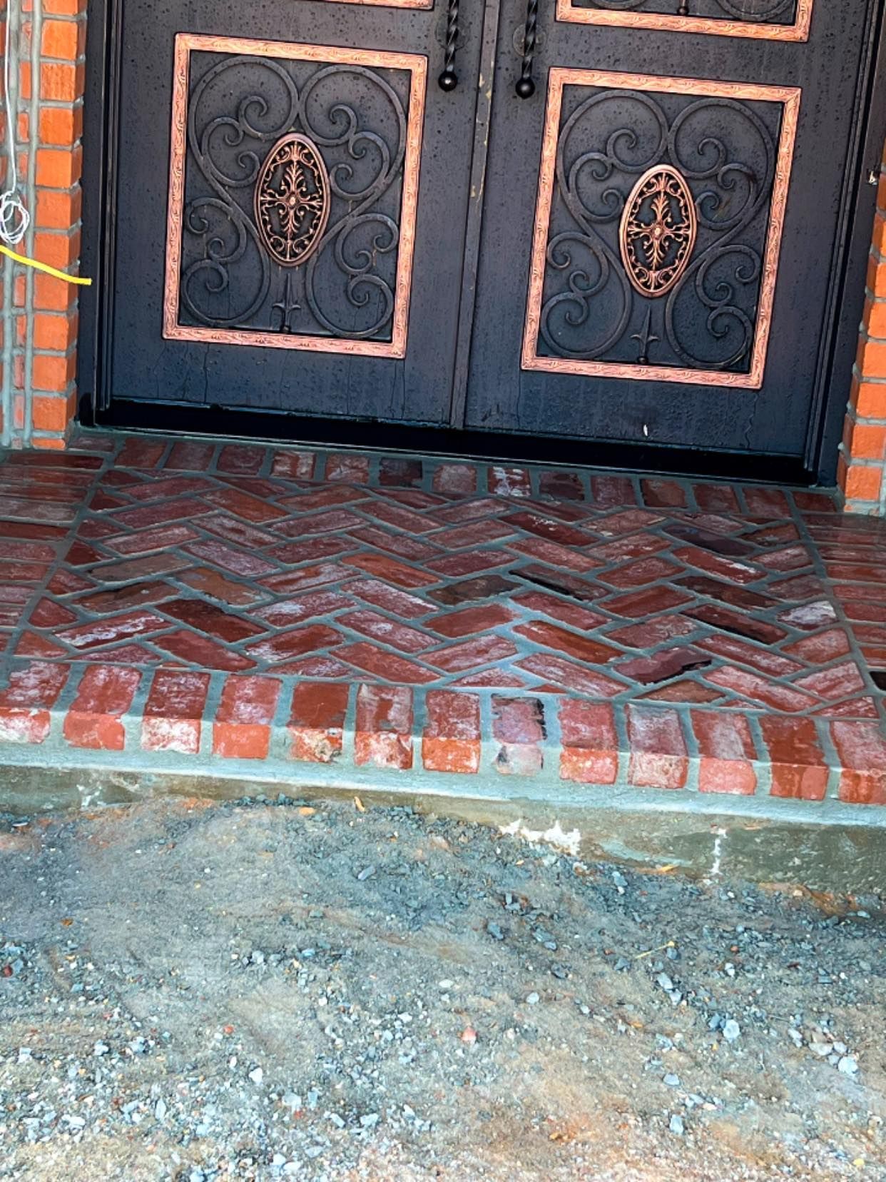 A black double door with decorative iron panels sits above a red brick stoop laid in a herringbone pattern.