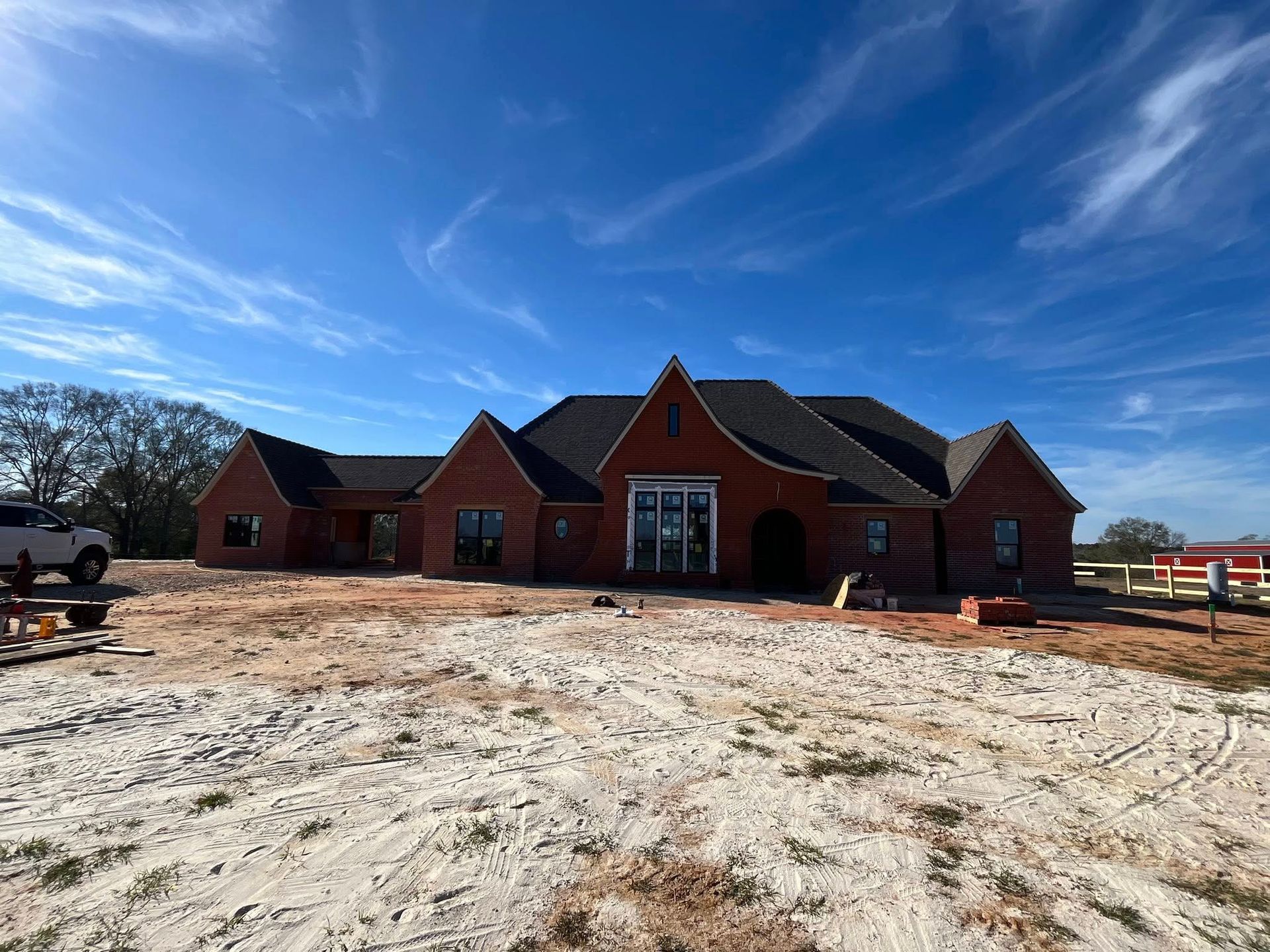 A single-story red brick house under construction, with a dark roof and large windows, sits on a sandy, bare dirt lot.
