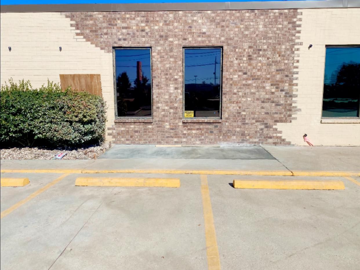 A building exterior with a tan wall partially covered by brick veneer, two central windows, and a paved parking lot.