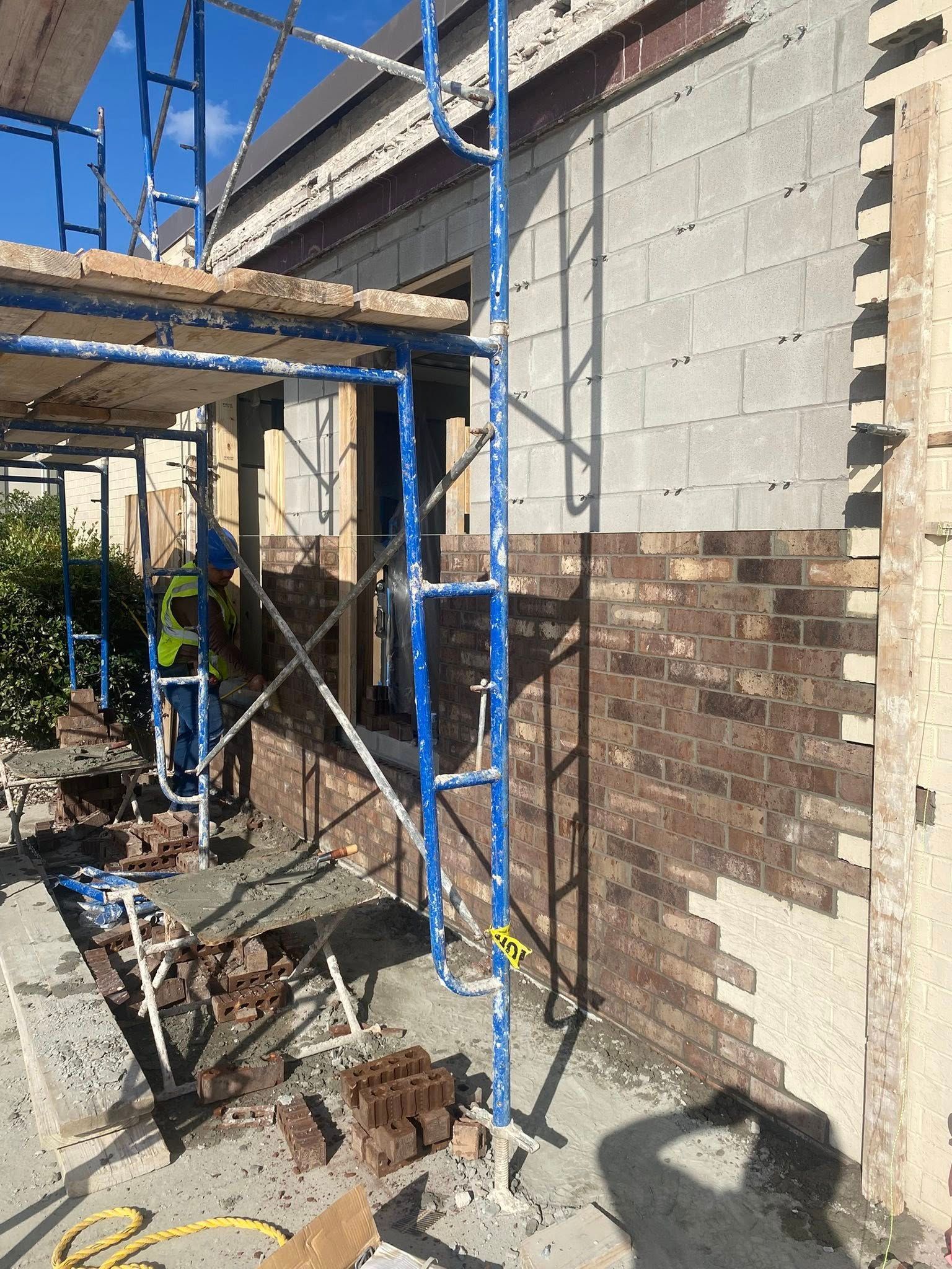 A construction worker in a high-visibility vest installs dark brick veneer on a block wall partially covered by scaffolding.