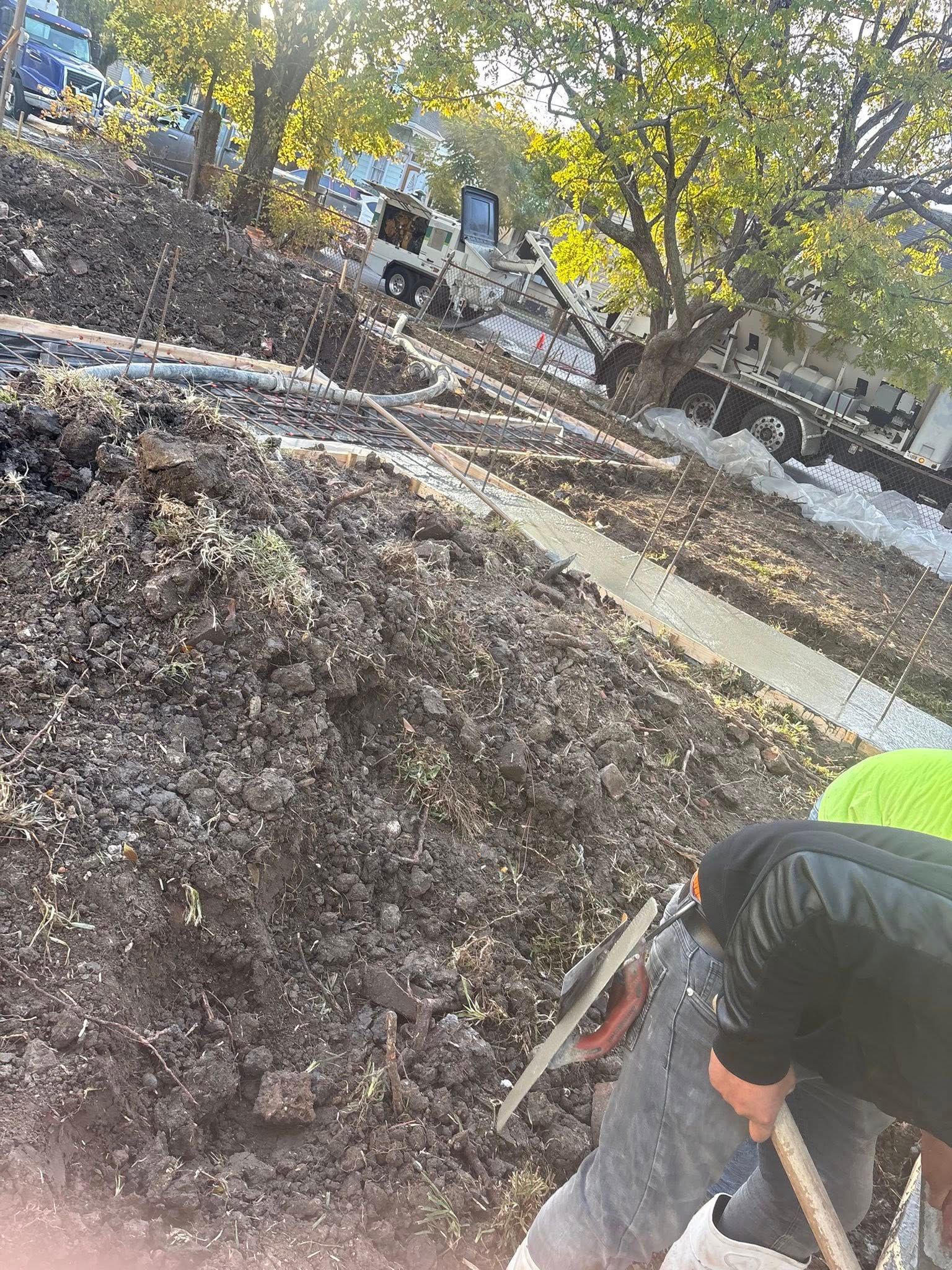 A worker in a high-visibility vest uses a hand tool on a large pile of dirt at a construction site with trees in the back.