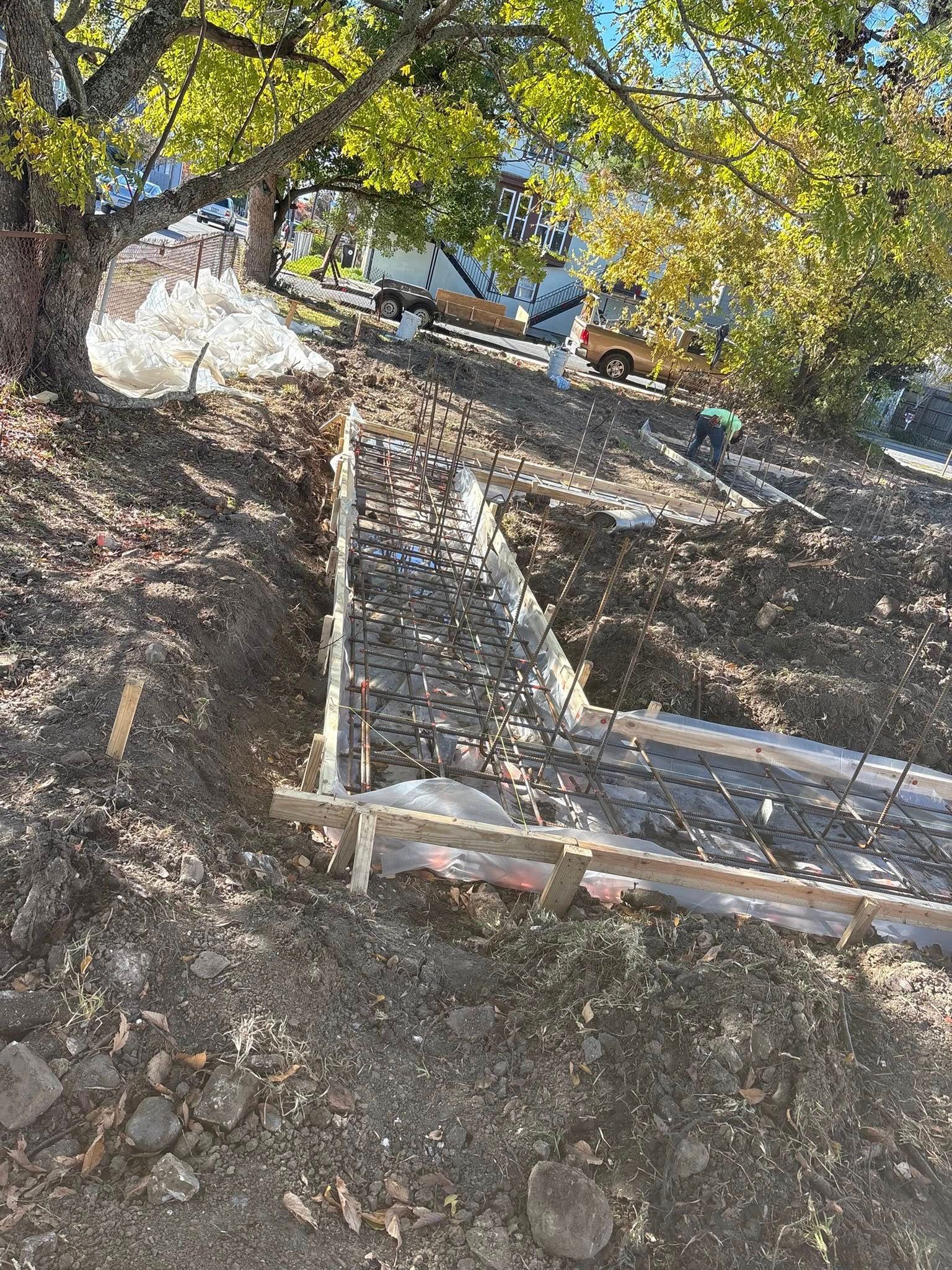 A wooden formwork frame is set in a dirt trench, prepared for a concrete foundation, beneath tree branches.
