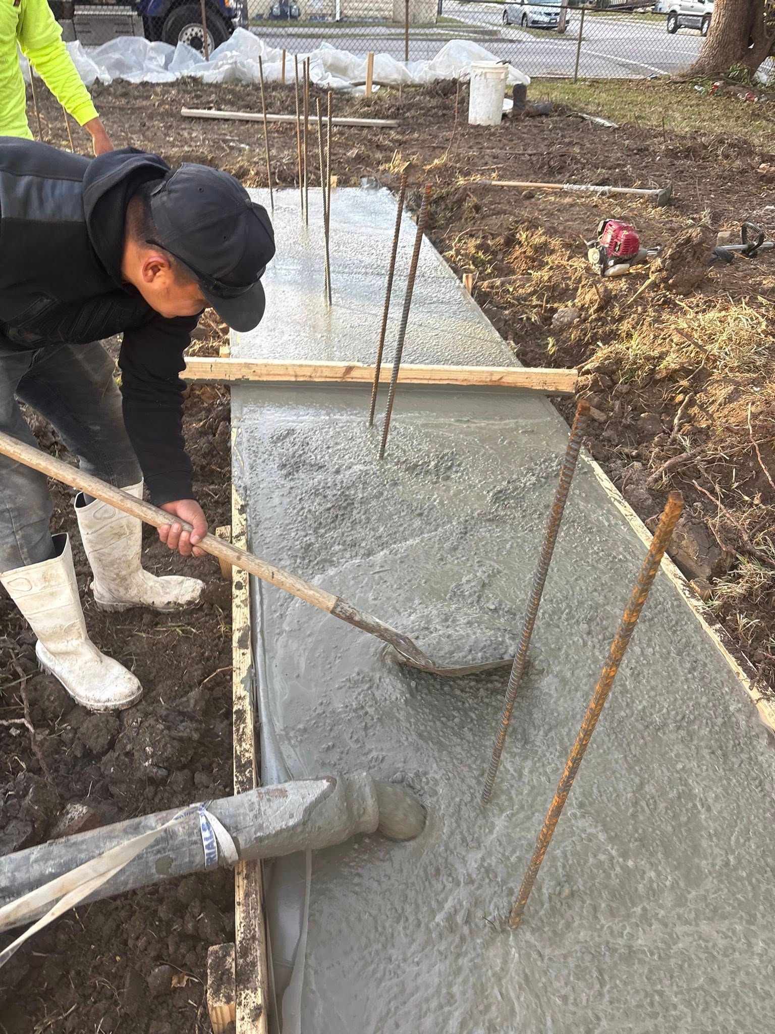 A person in a black jacket and white boots uses a rake to level wet concrete poured into a wooden foundation form.