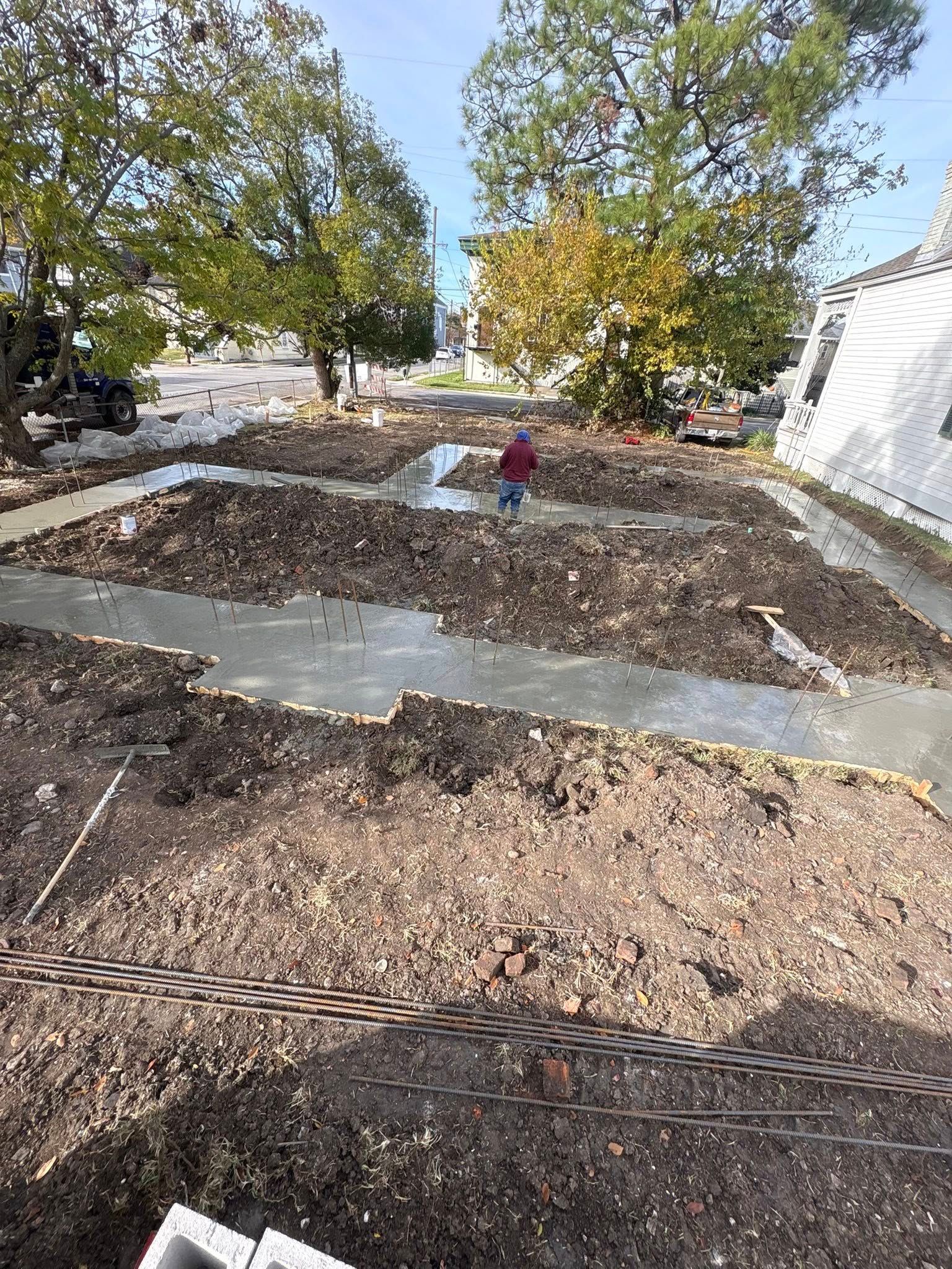 Fresh concrete foundations are poured for a construction project in a dirt-filled lot near a white building and trees.